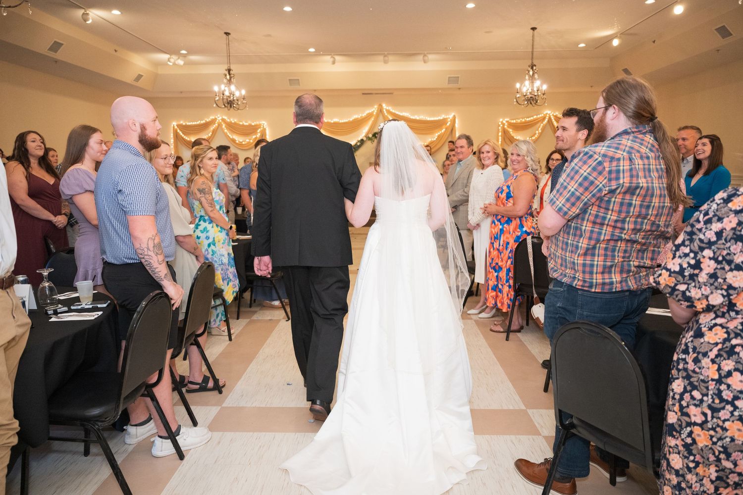 Wedding ceremony in progress inside an elegant ballroom with guests standing and chandeliers overhead. Pease Minnesota Wedding at Northern L