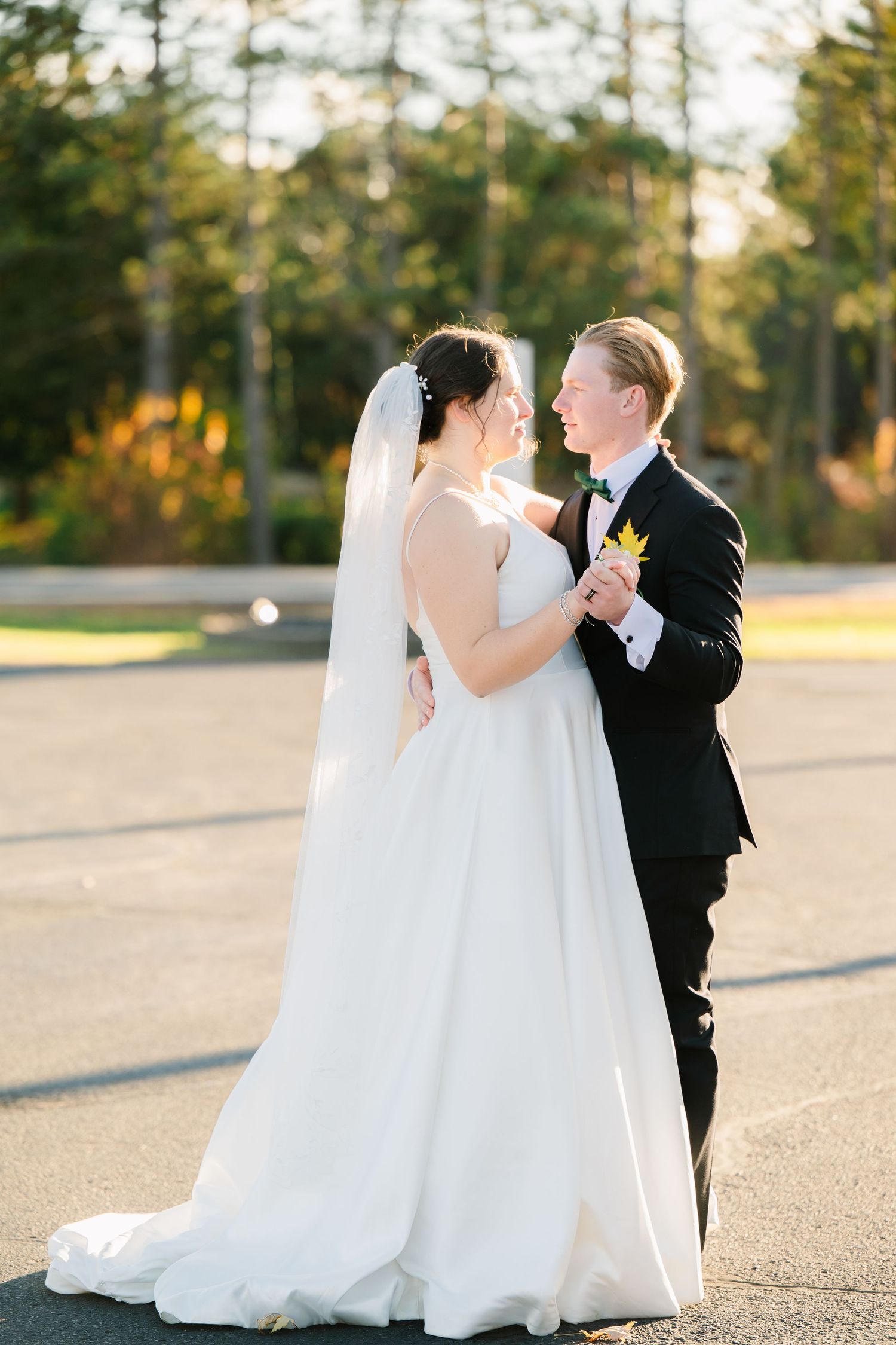 Wedding couple shares intimate moments in formal attire on a sunlit paved area surrounded by evergreen trees.