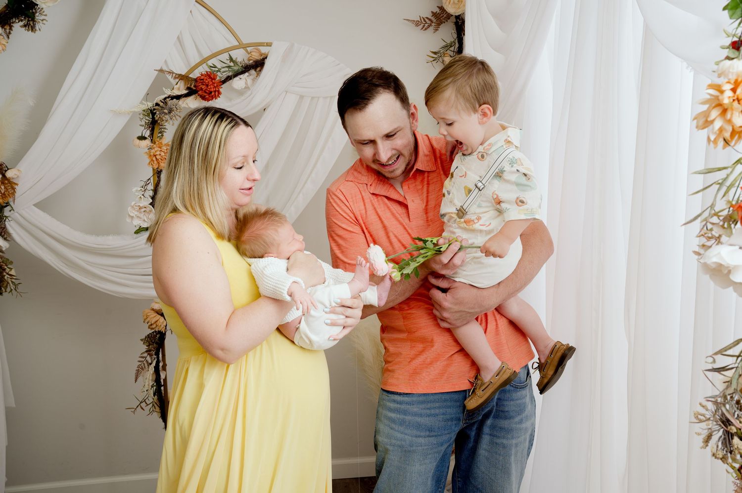 Family stands together in front of white drapes with floral decor while holding their newborn baby in yellow dress.
