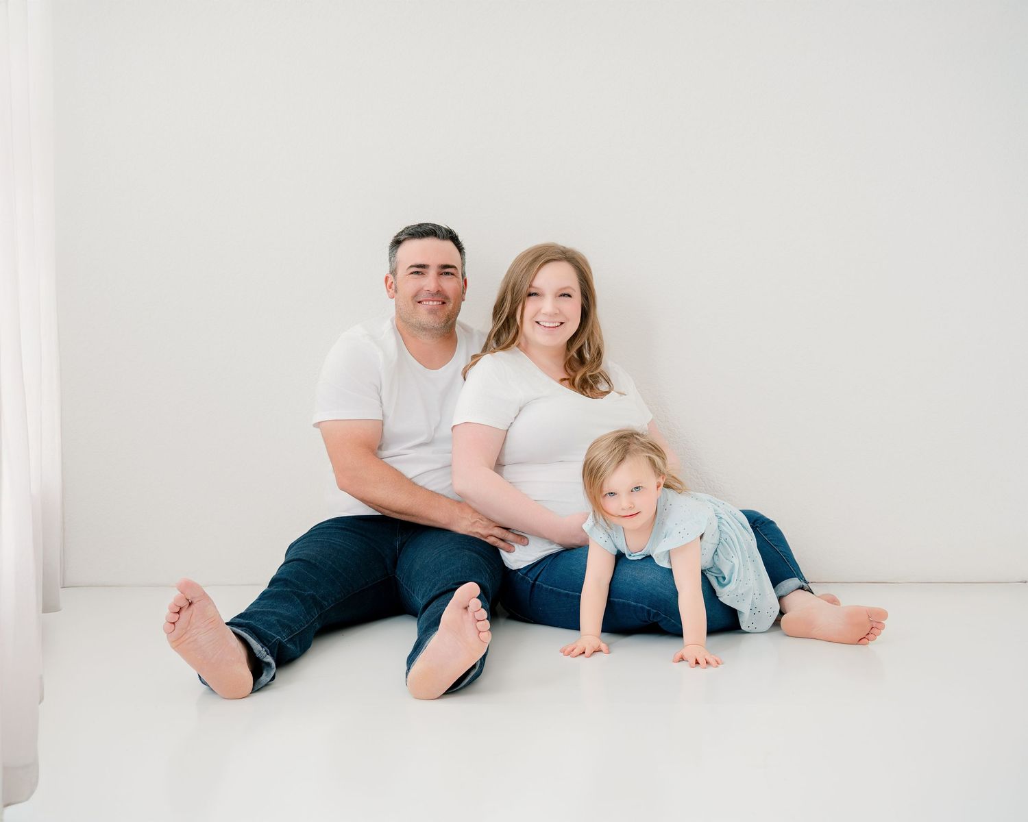 A family of three poses together on a white floor for a casual indoor portrait session.