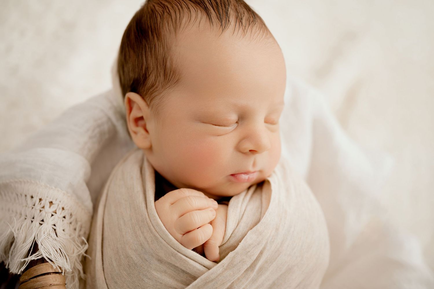 Peaceful sleeping newborn wrapped in soft white swaddle against light background.