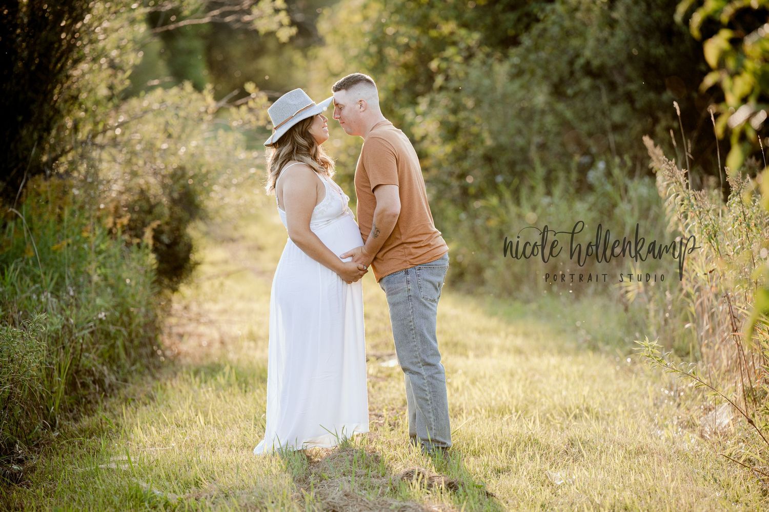 A couple in a sunlit meadow embraces during a maternity photoshoot at golden hour.
