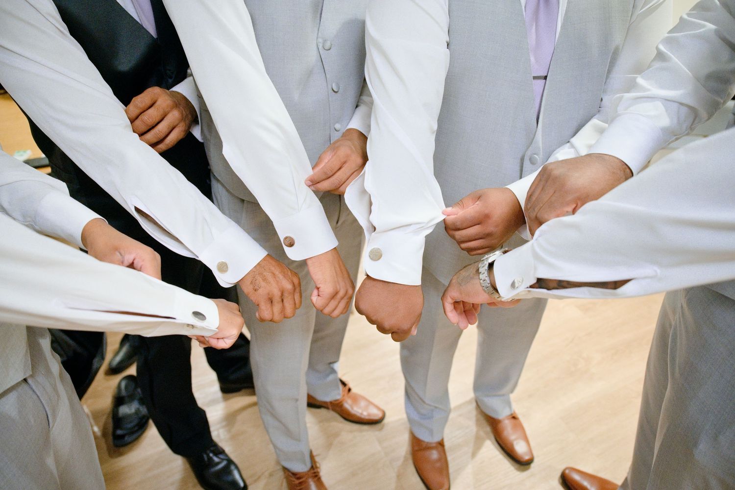 Groomsmen in matching light gray suits adjusting cufflinks together before a wedding ceremony.