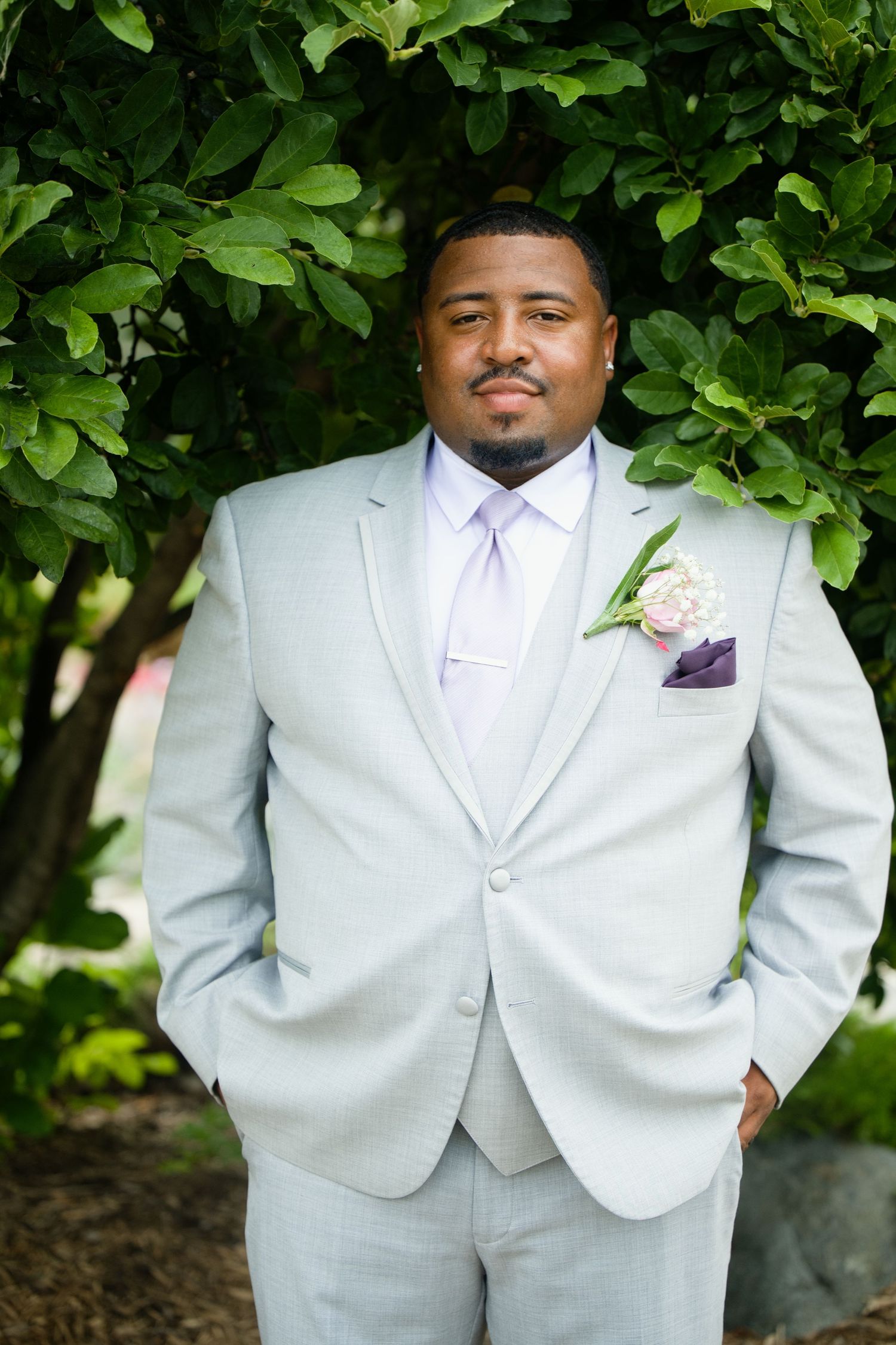A groom in a light gray suit with lavender tie and floral boutonniere poses outdoors before his wedding ceremony.