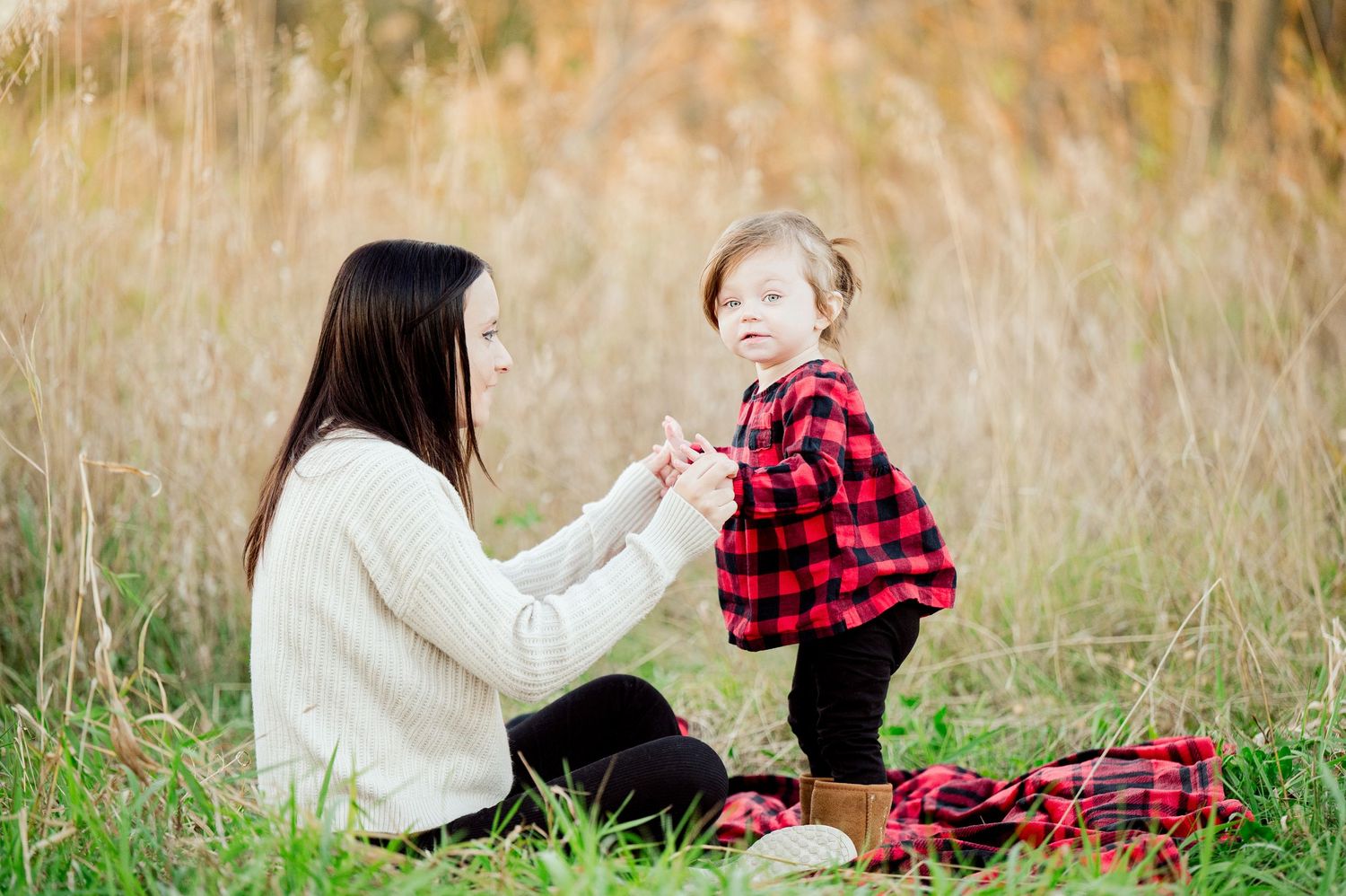 Two people sit in a tall grass field wearing red plaid and cream colored sweaters during an autumn photoshoot.