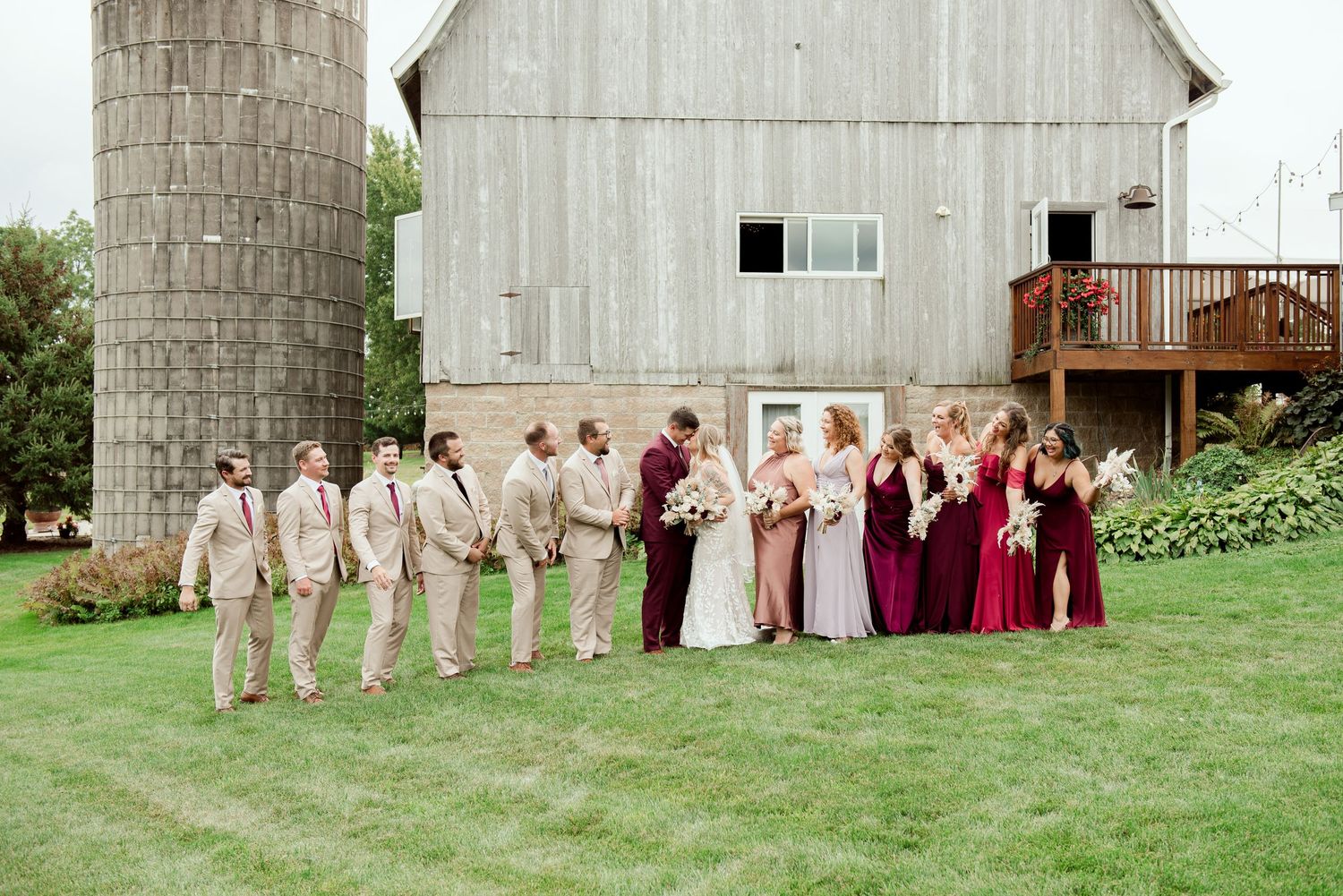 Wedding party poses in front of rustic white barn with silo in formal attire.