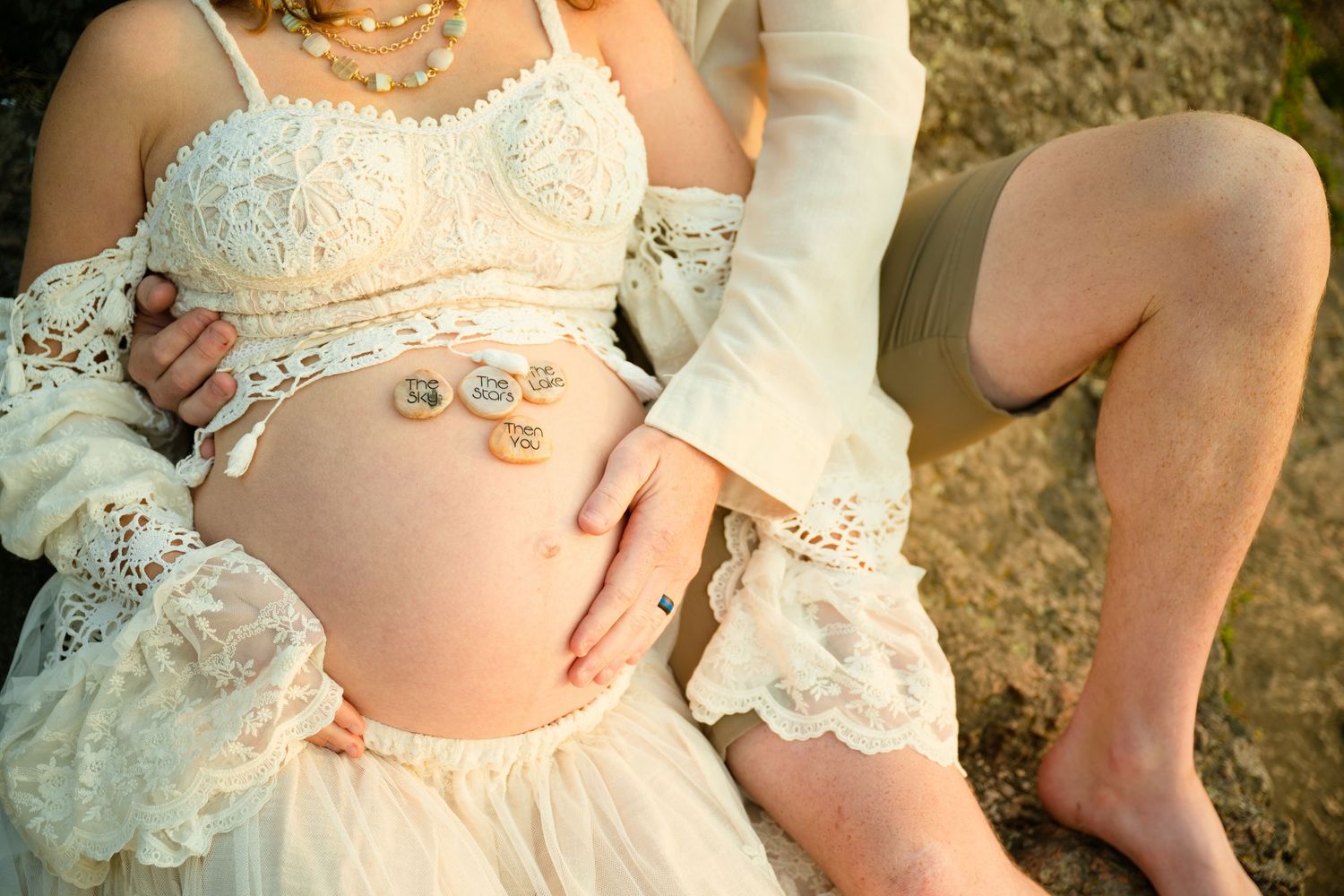 Pregnant woman in lace boho dress with partner's hand on baby bump, outdoor maternity photo session.