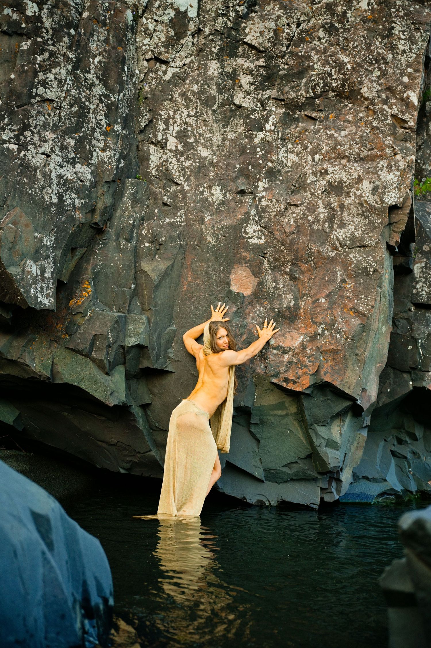 Woman in sheer dress standing in water against dramatic rocky cliff wall in a serene natural setting.