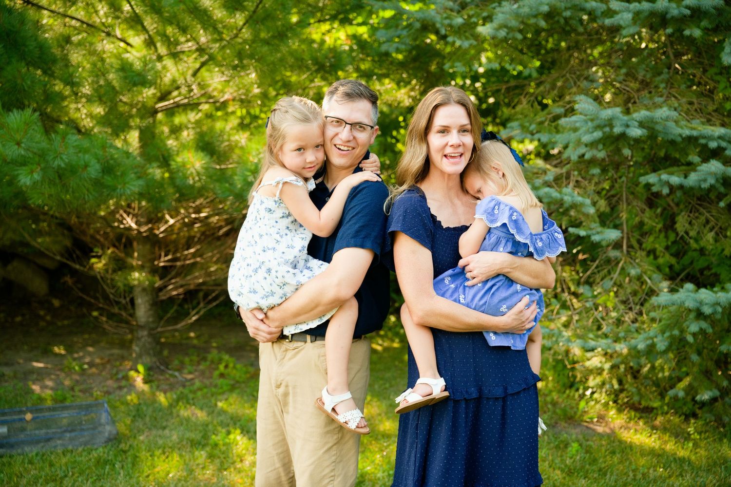 A happy family of four poses outdoors, with parents holding two young children amid lush green trees.