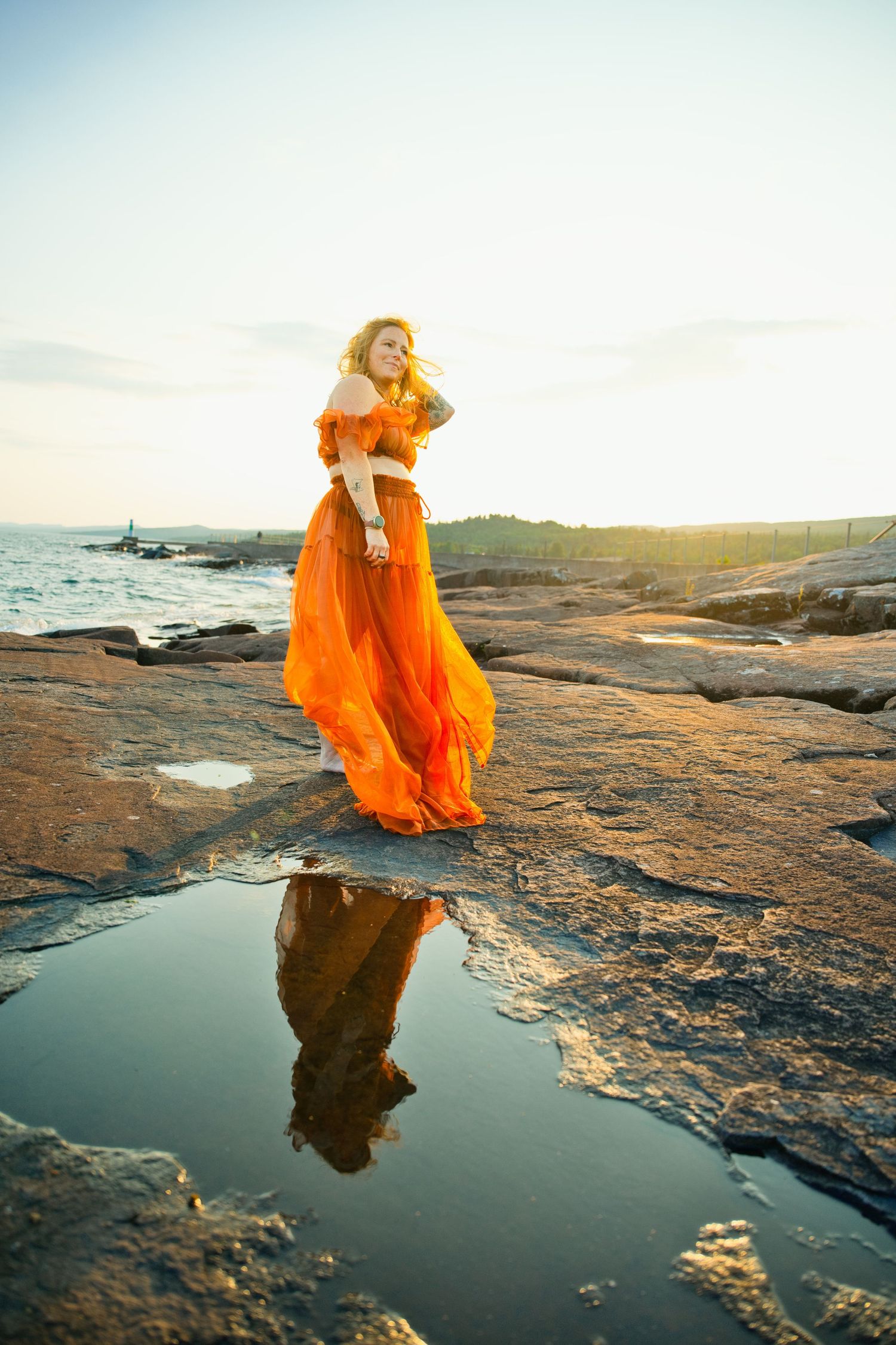 Woman in flowing orange dress stands on rocky coast with reflection in tide pool at golden hour.