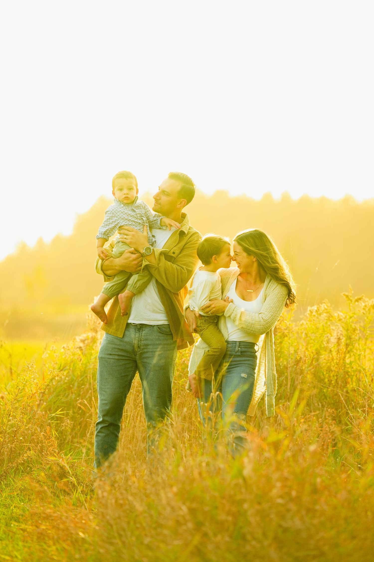 A loving family of four with two young children stands together in a golden sunlit meadow at sunset.