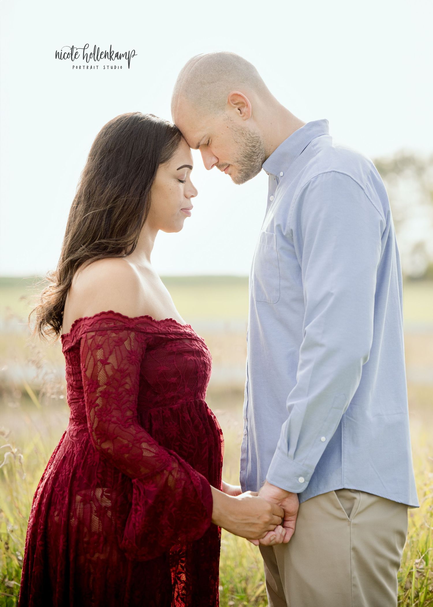 Couple in maternity photo shoot, woman in red lace gown and man in blue shirt holding hands in a field.