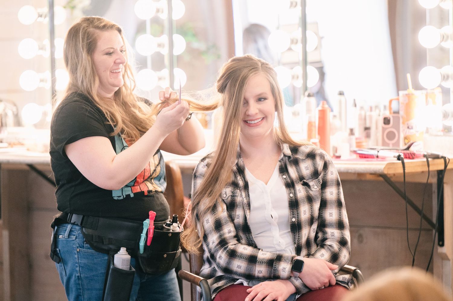 A hairstylist styling a young woman's long hair in a bright salon with vanity mirrors.