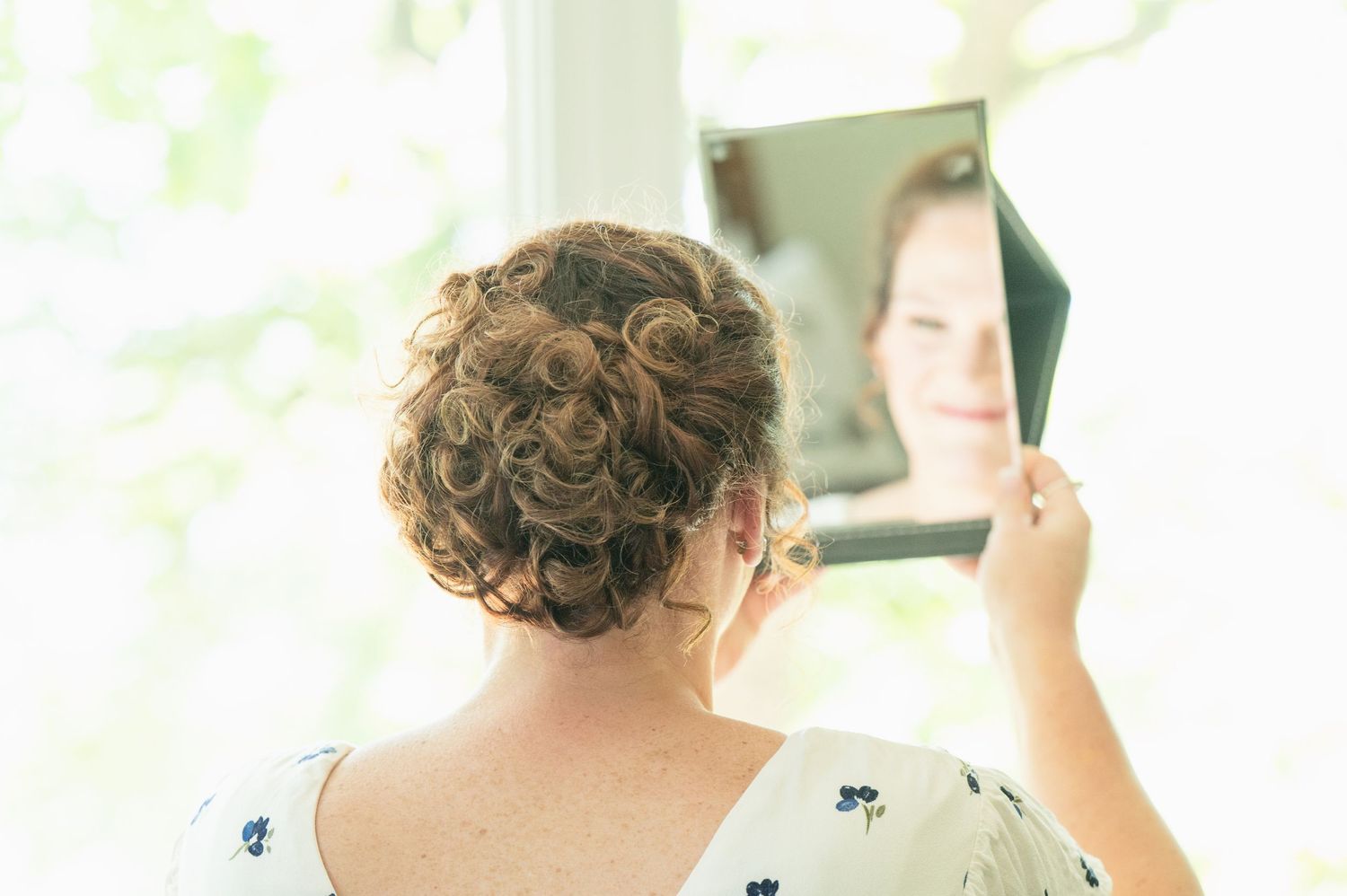 Woman with curly updo hairstyle holding a mirror, smiling at her reflection in soft natural light.