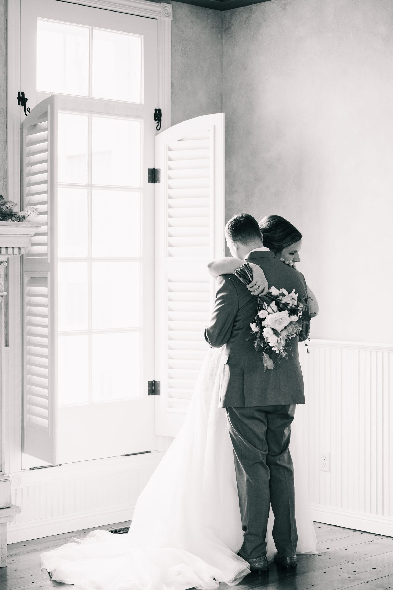 Black and white photo of bride and groom sharing first look embrace near white louvered doors.