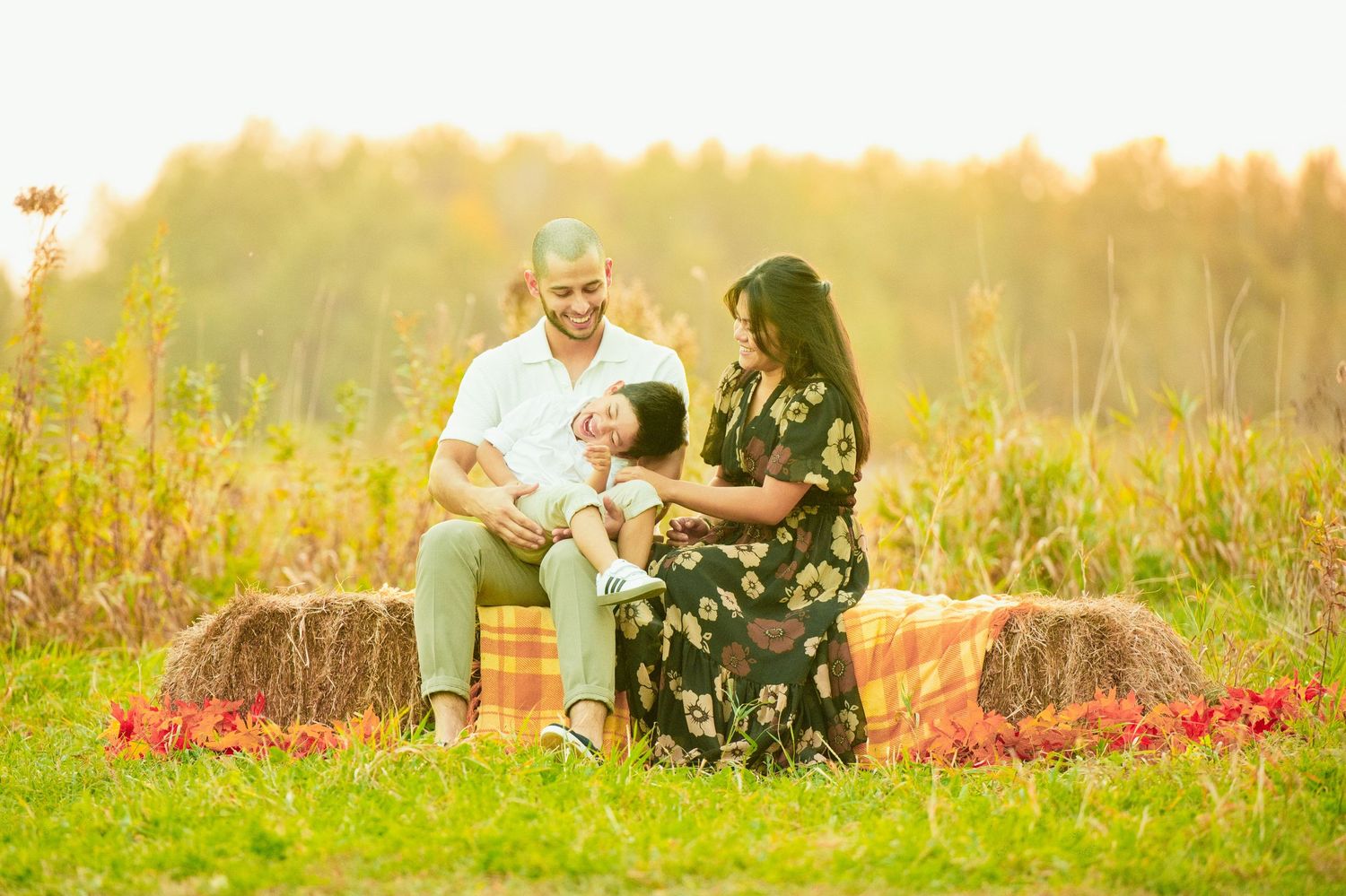 Family of three enjoying a sunny outdoor session on hay bales in a golden field, parents smiling at their baby.