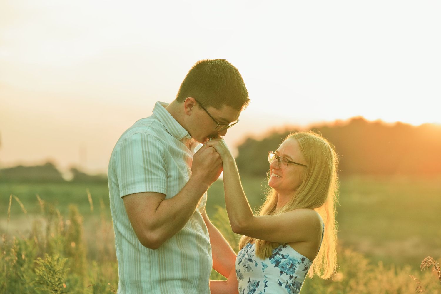 Romantic couple holding hands in a sunny golden field at sunset, man kissing woman's hand outdoors.