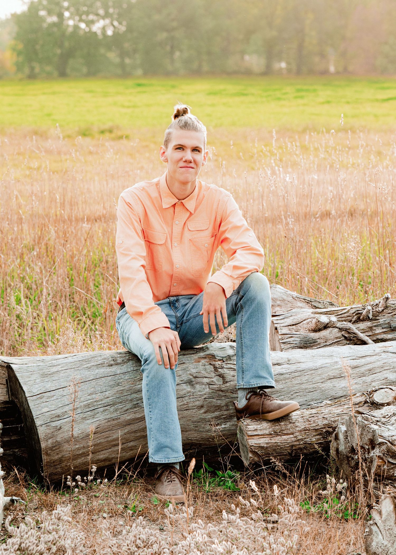 Person in coral shirt and jeans sitting casually on fallen log in golden field during sunset.