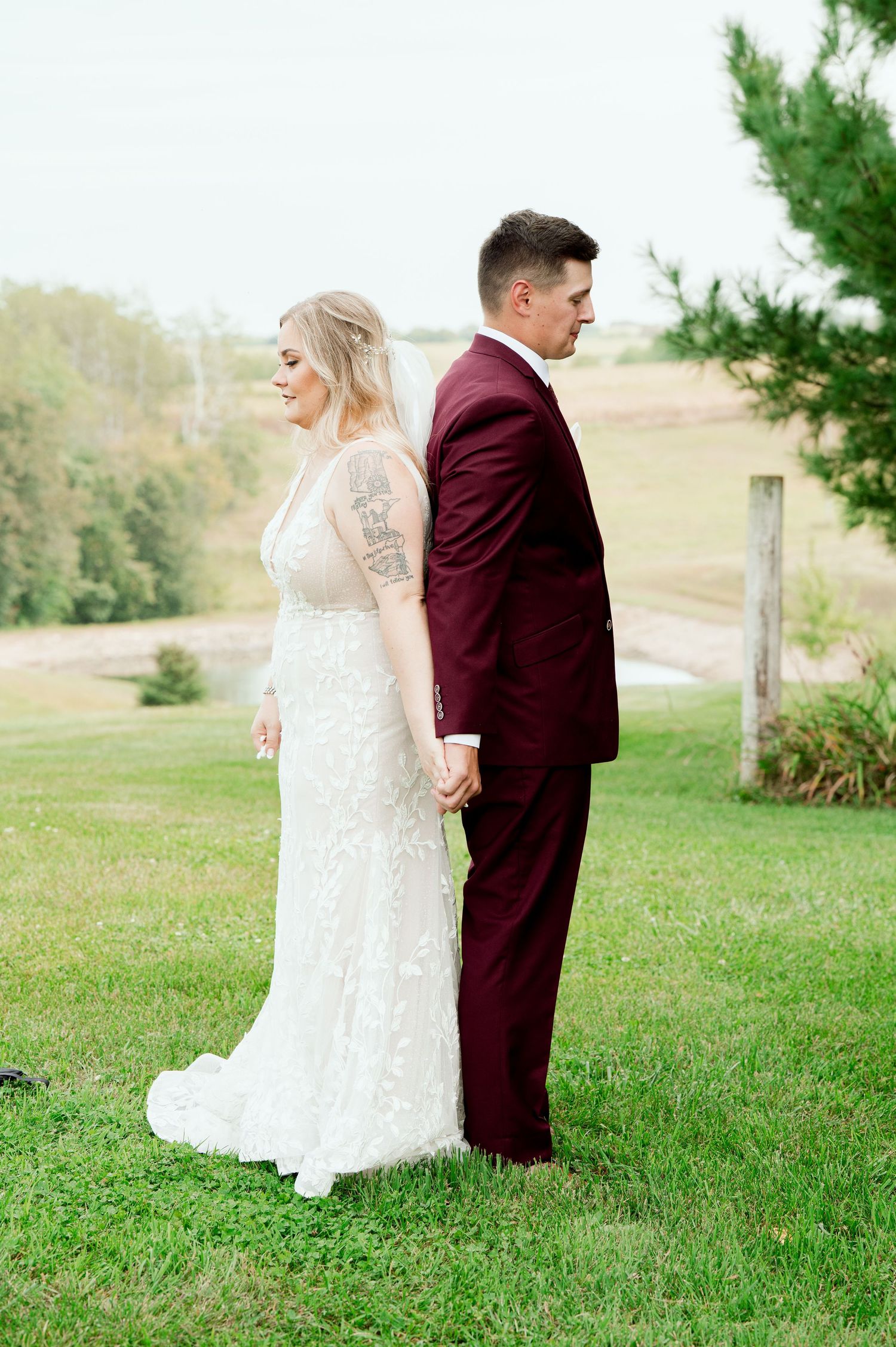 A couple stands back-to-back on a grassy hill, with the bride in white lace and groom in burgundy suit holding hands.