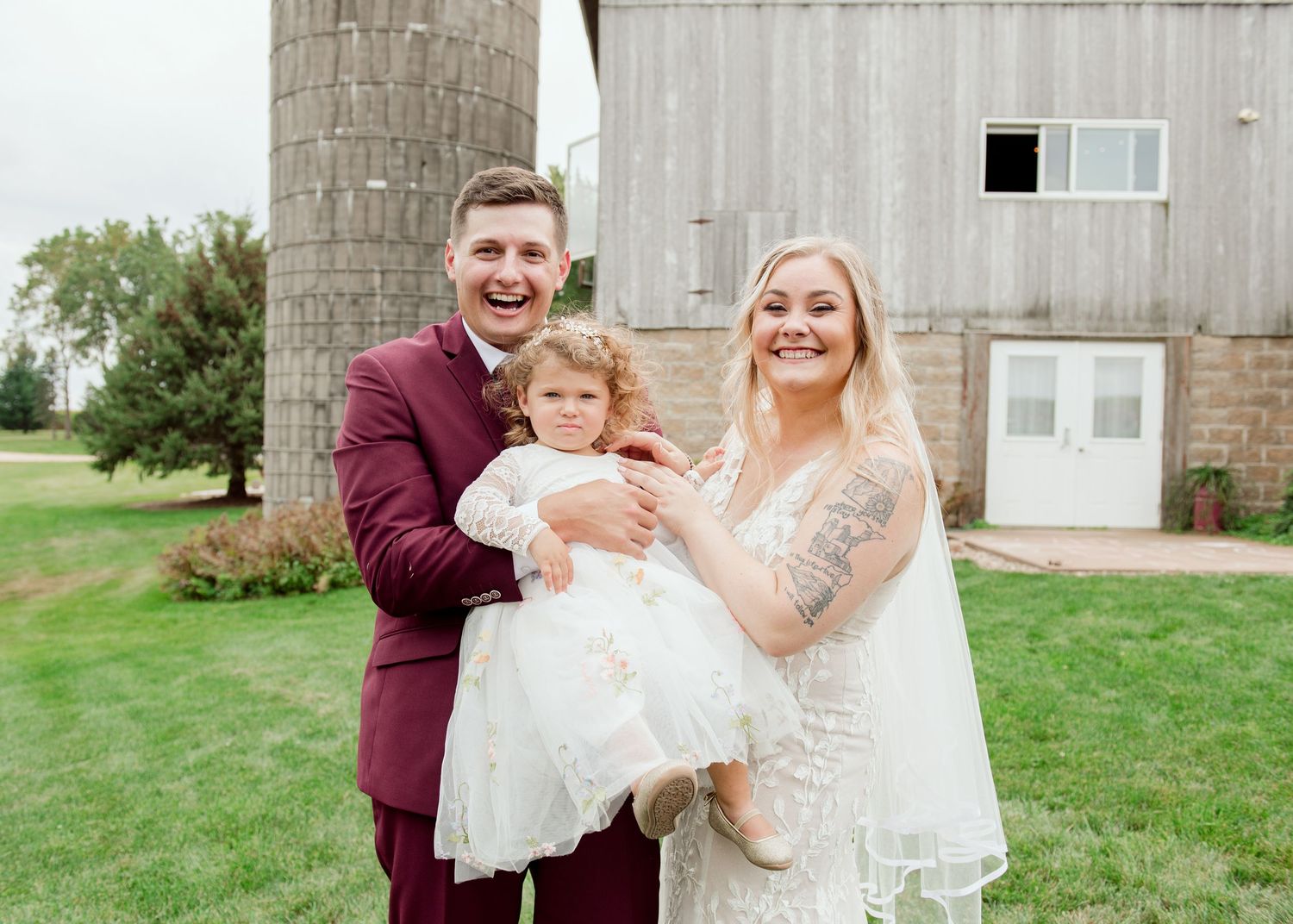 Family portrait with toddler in white dress against rustic barn backdrop.