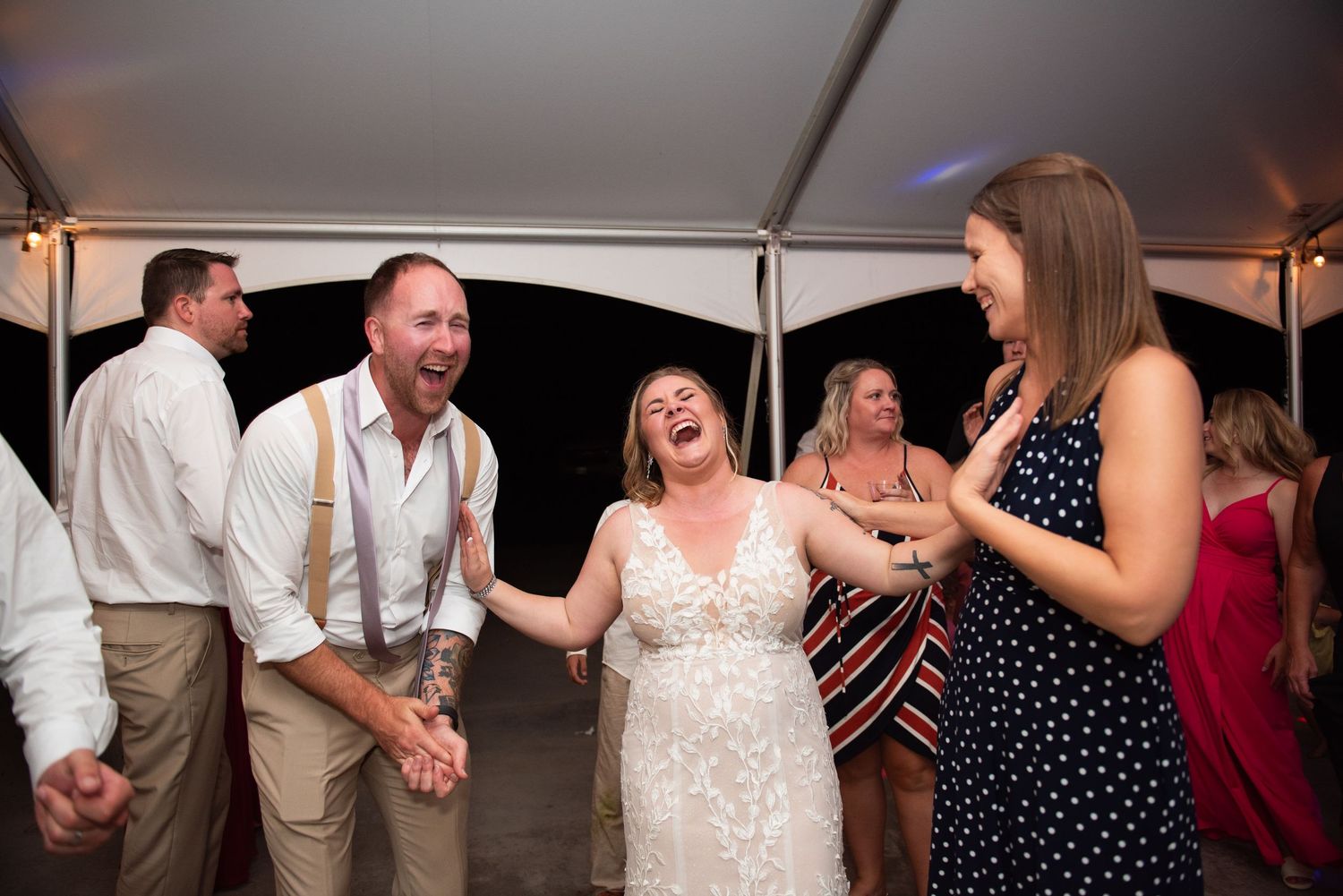 Wedding guests dancing and laughing together during the reception under a tent with festive lighting.