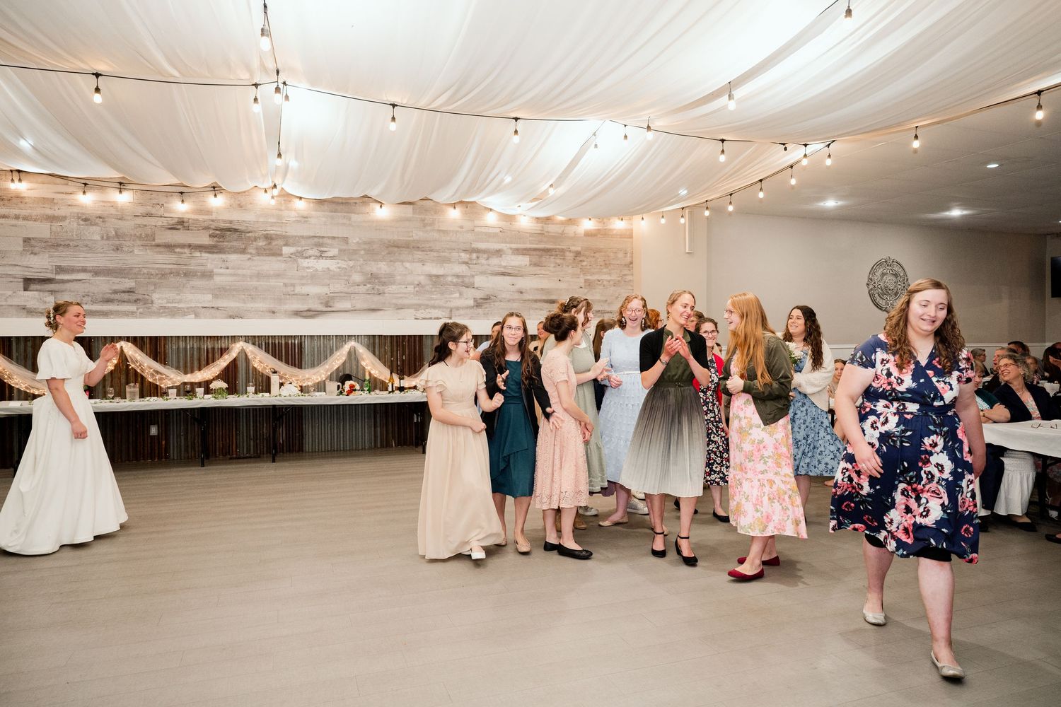 Wedding reception guests dancing and celebrating in a decorated indoor venue with string lights.