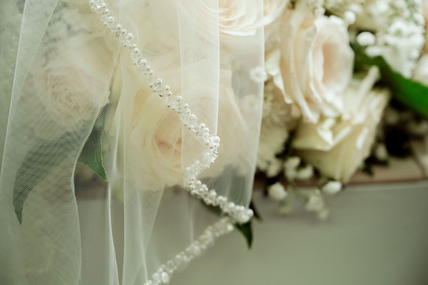 White veil with delicate pearl trim lies beside soft pink roses and baby's breath in an elegant bridal arrangement.