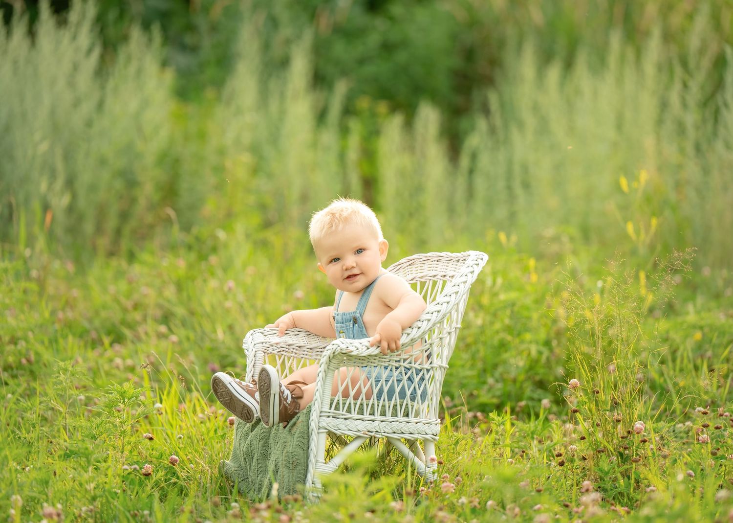 A smiling baby in a teal romper sits in a white wicker chair surrounded by lush green meadow grass outdoors. Milestone and Birthday Photogra