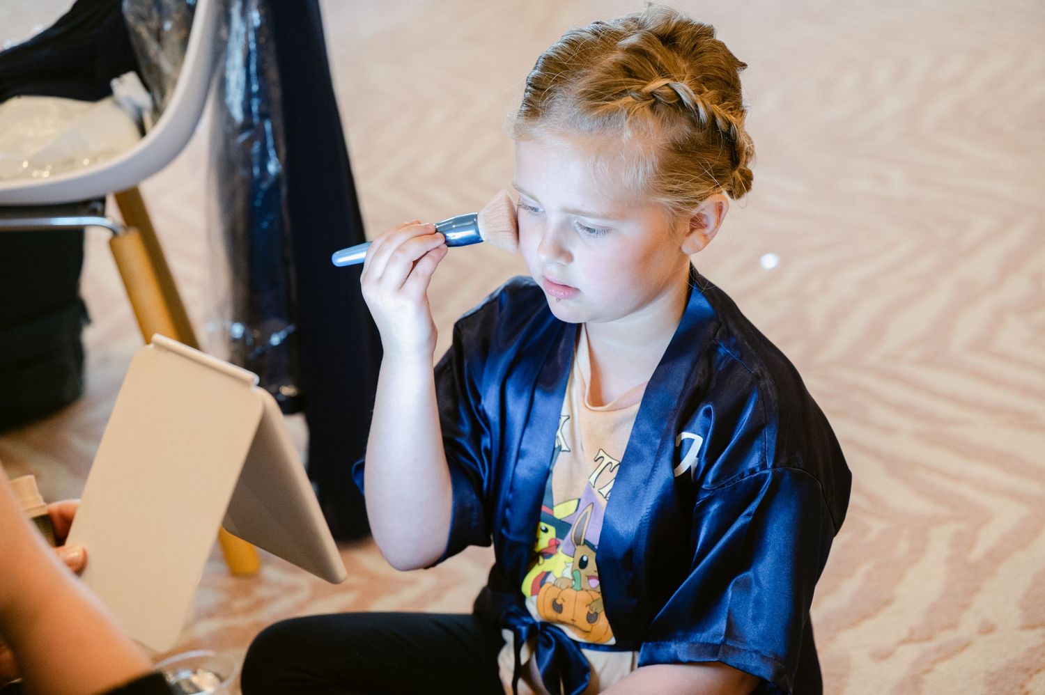 Central MN Wedding Photos | Young flower girl in blue satin robe applies makeup using a small mirror during wedding preparations.