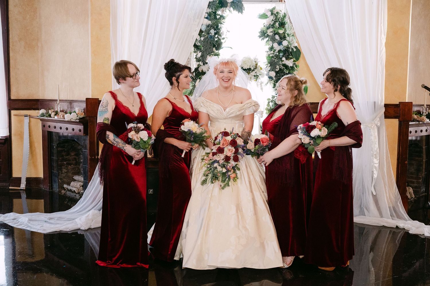 Central MN Wedding Photos | Bride in off-shoulder ivory ballgown stands with four bridesmaids in burgundy velvet dresses holding bouquets.