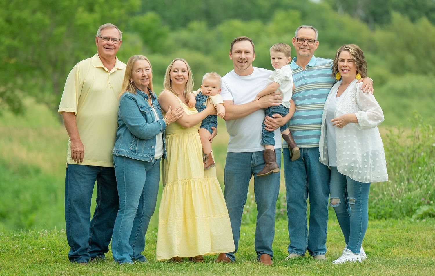 A multi-generational family stands together in a grassy field wearing casual clothing on a sunny day.