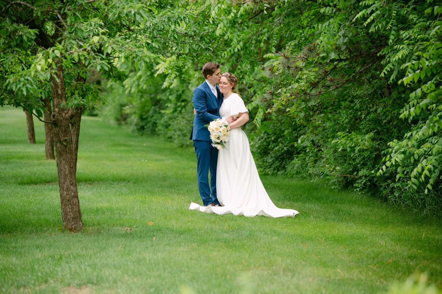 A romantic wedding couple embraces on a lush green lawn surrounded by trees and natural scenery.