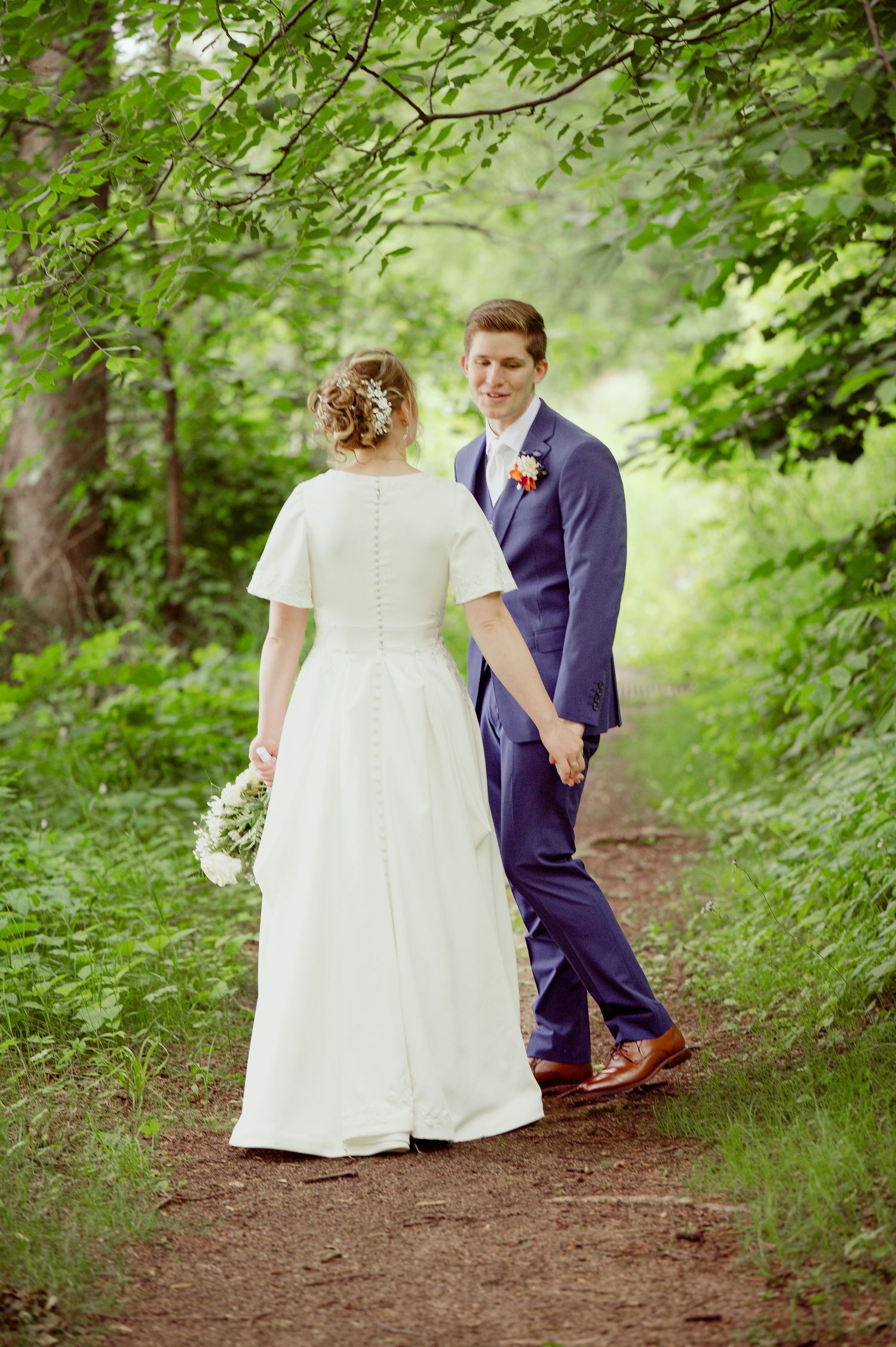 A couple in wedding attire walks along a wooded dirt path surrounded by greenery.