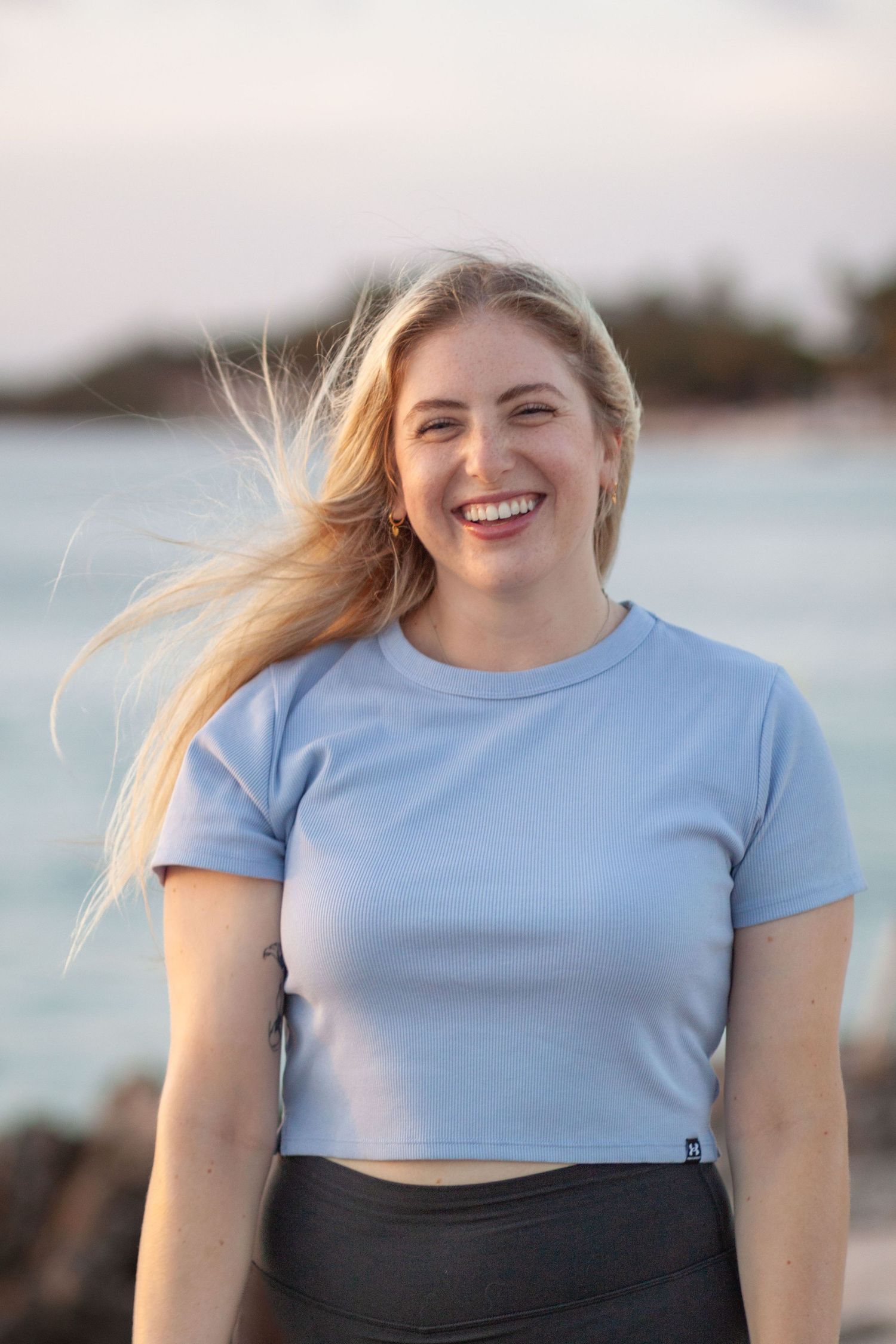 Woman in blue t-shirt smiles brightly outdoors by the water.