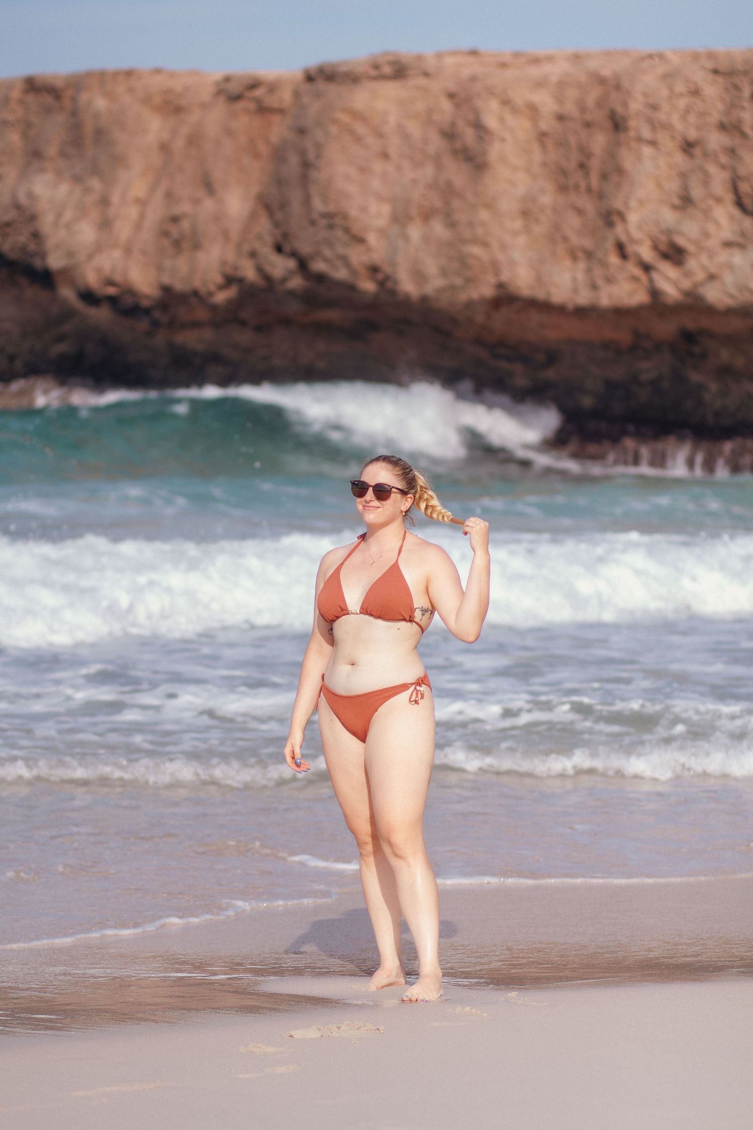 A woman in a red bikini enjoys a sunny day at a scenic beach with rocky cliffs in the background.