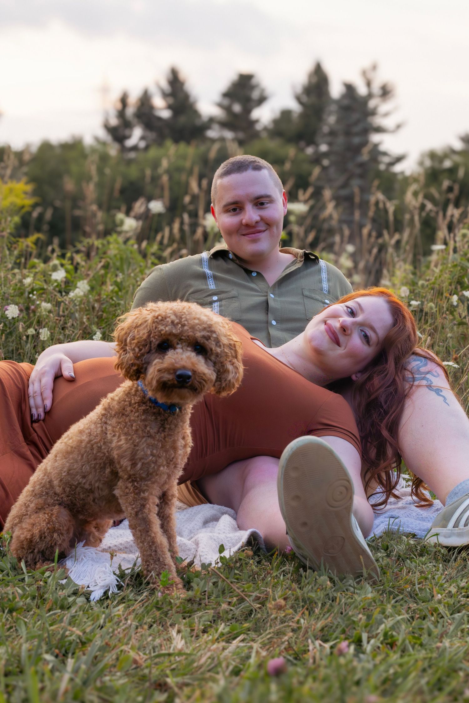 A relaxed maternity photo features a couple lying in the grass with their curly-haired dog nearby.