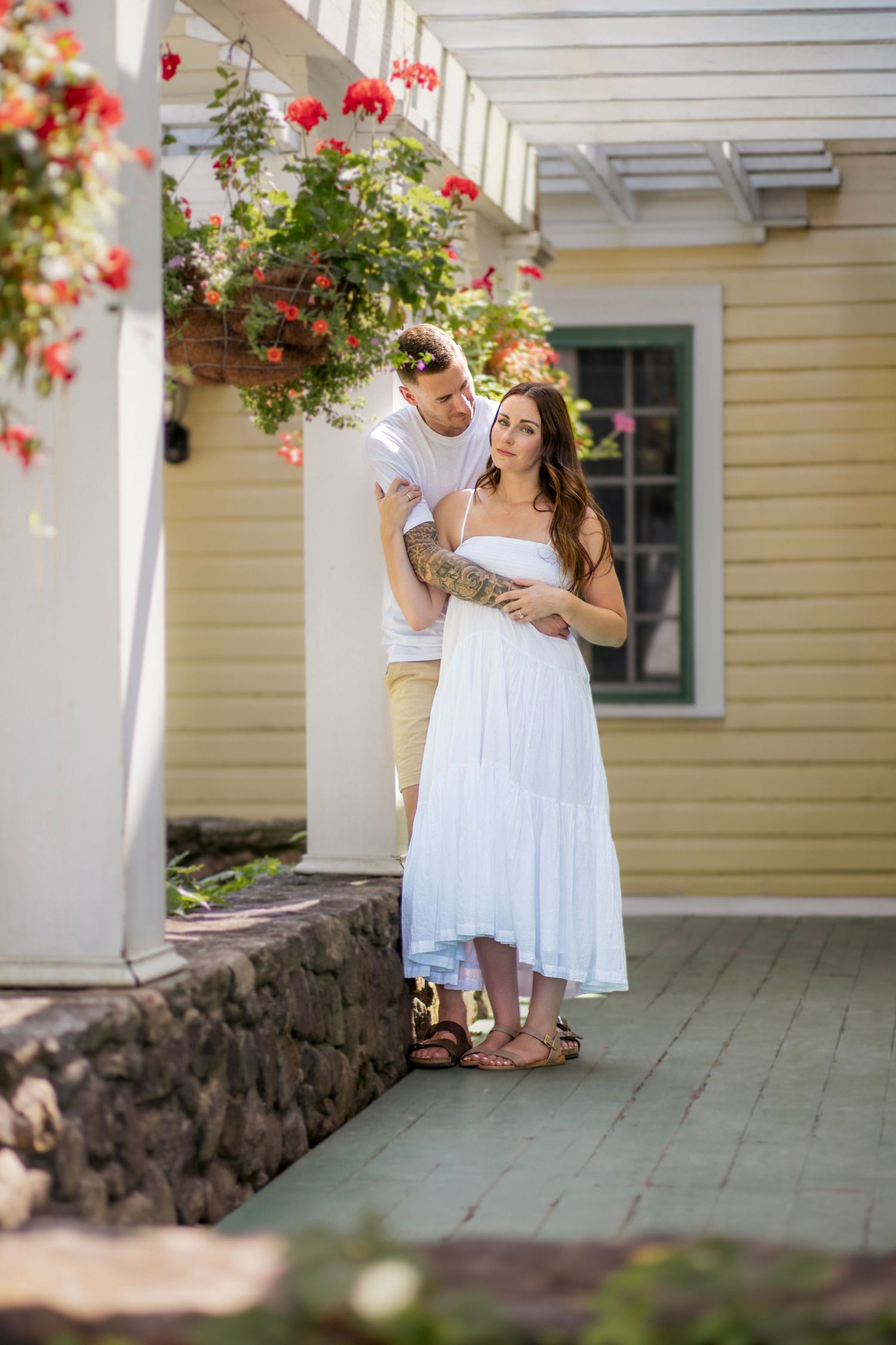Couple shares intimate moment on porch with red flowers and stone wall in background.