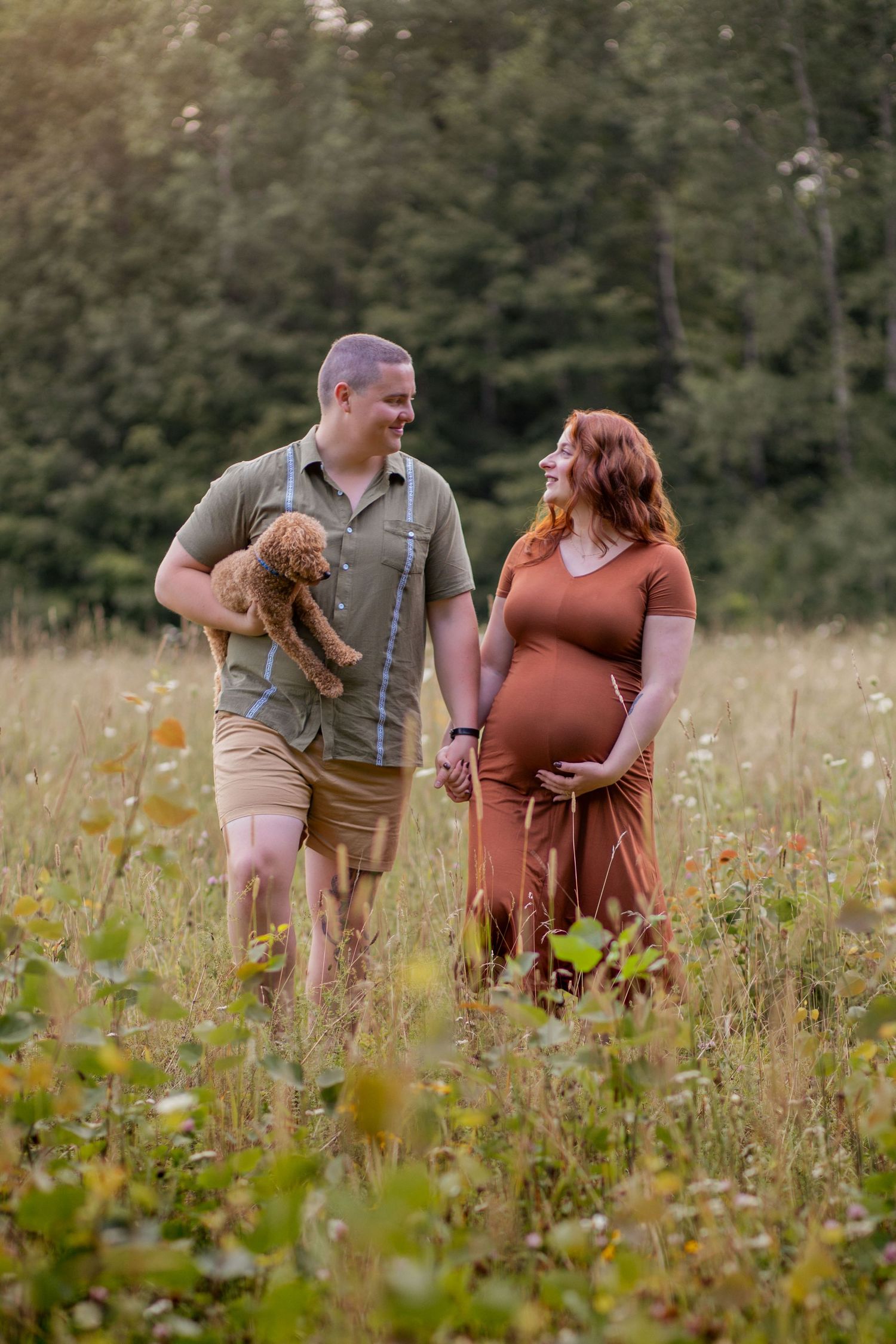 A couple walks hand in hand through a summer meadow during their maternity photoshoot.