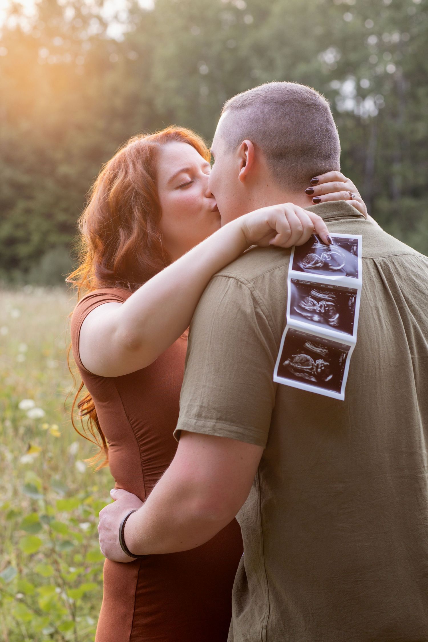 Romantic couple holds ultrasound image while embracing in sunlit meadow.