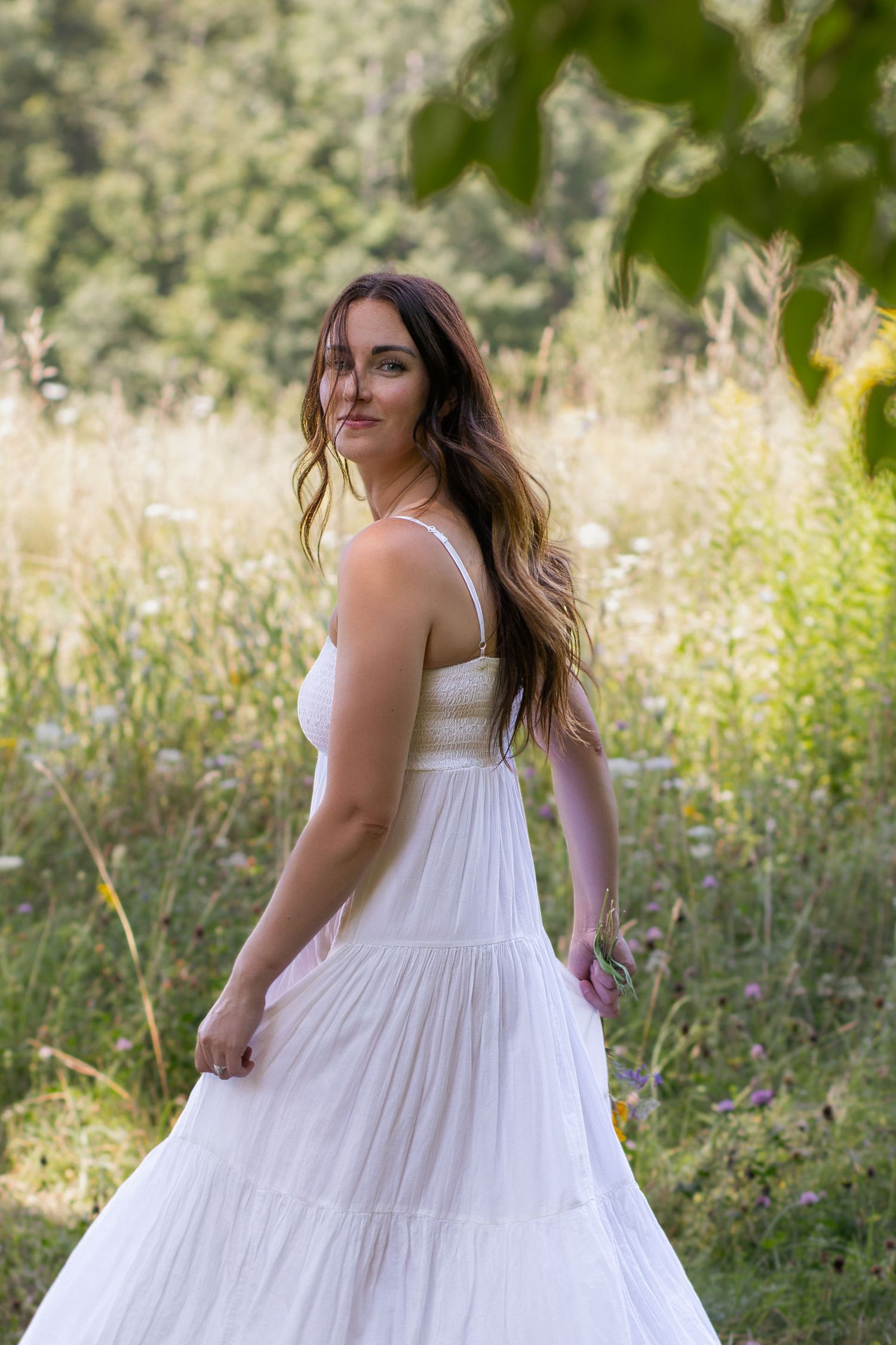Woman in flowing white dress poses naturally in a sunlit summer field.