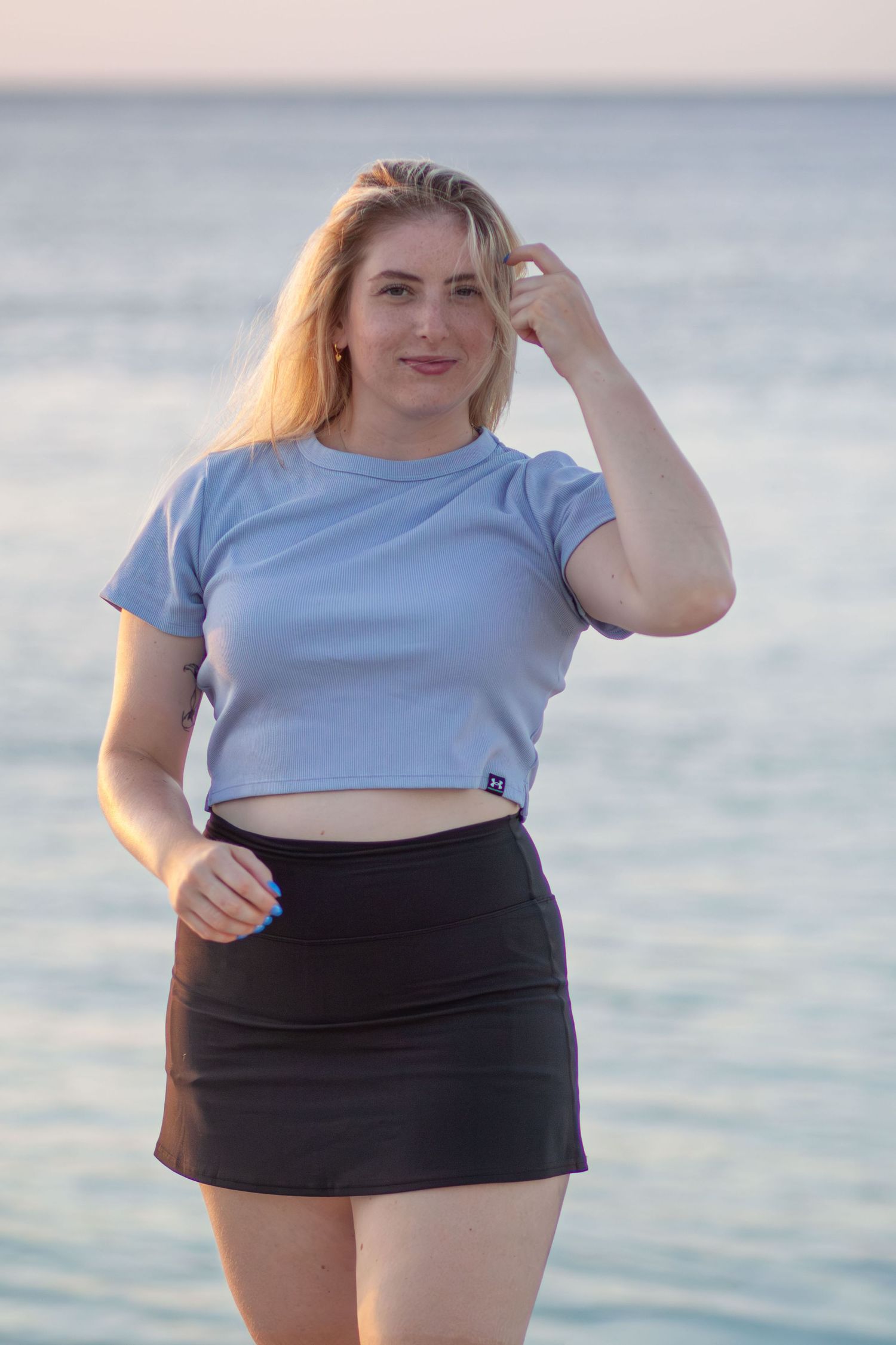 A person in a light blue crop top and black skirt poses confidently near a waterfront on a sunny day.