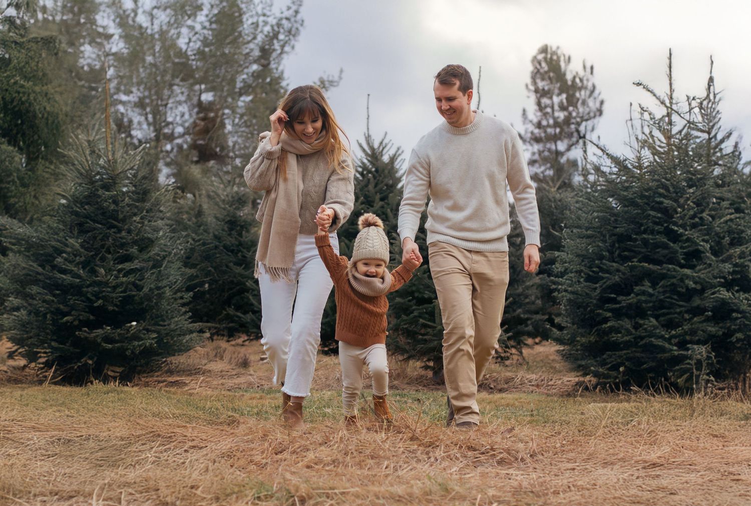 Couple strolls together through field with evergreen trees in background.