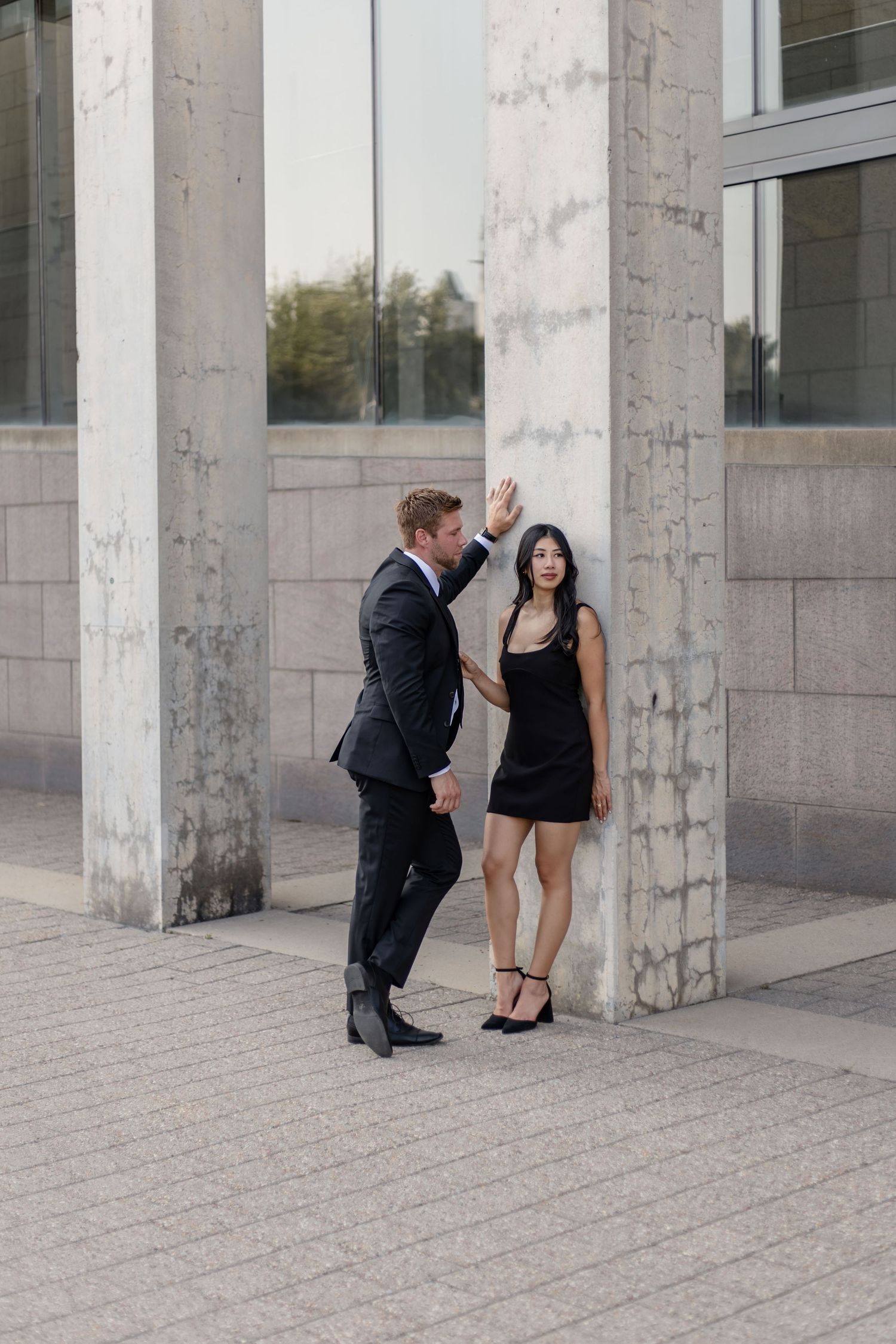 A couple in formal wear posing playfully between concrete columns.