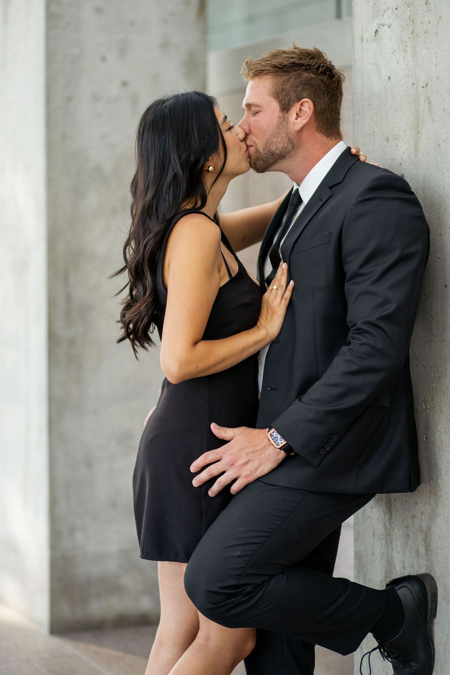 A couple in formal attire sharing an intimate moment against a concrete wall.