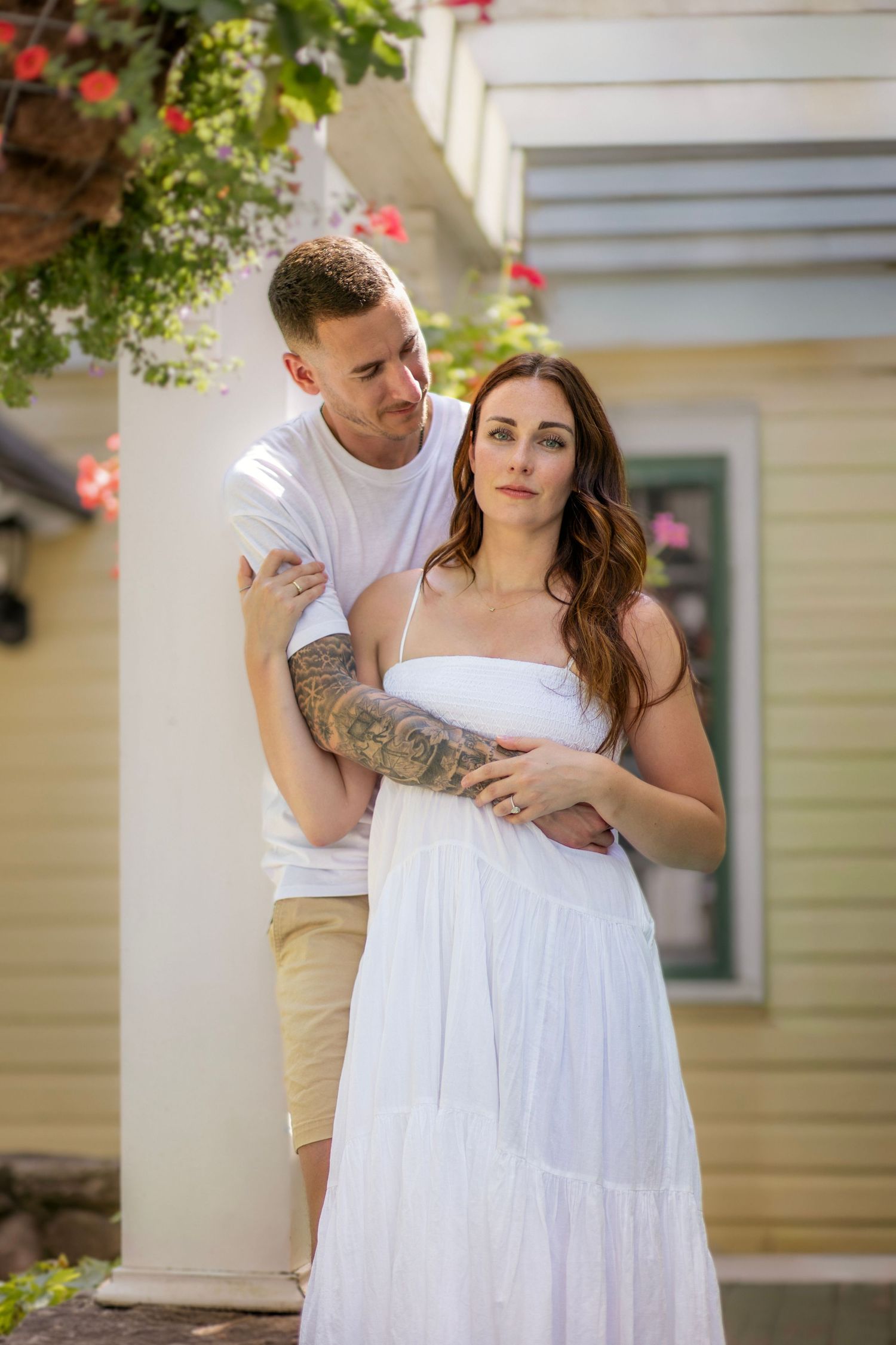 Couple shares tender moment on sunlit porch decorated with hanging red flower baskets.