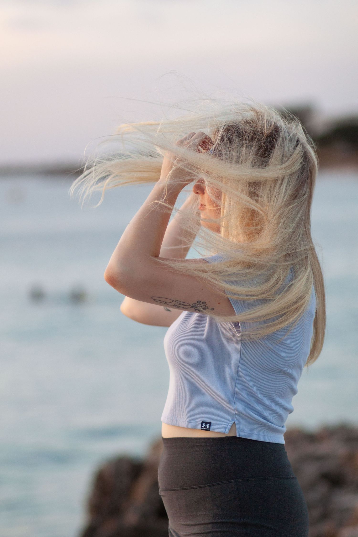 Woman in light blue top with blonde hair poses against seaside sunset with hair blowing in wind.