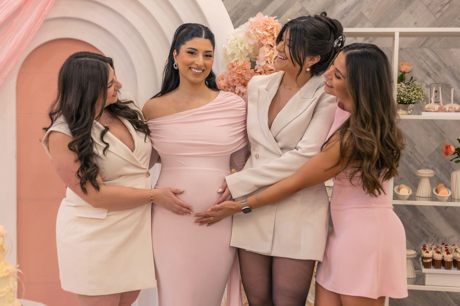 Four women at a baby shower, with the pregnant guest of honor in a pink gown as friends touch her baby bump.