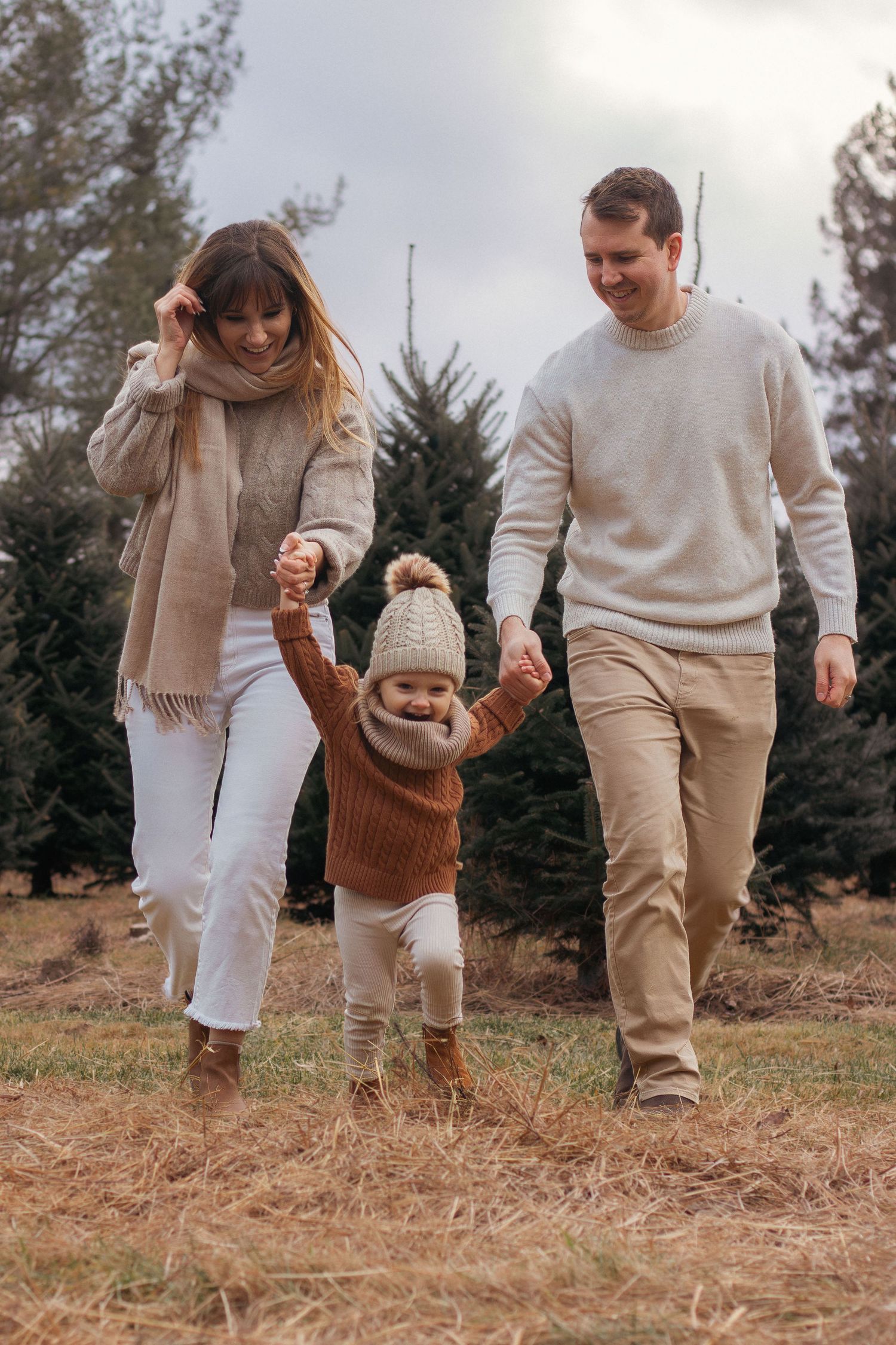 A couple walks through grass wearing matching cream sweaters and neutral tones near evergreen trees.