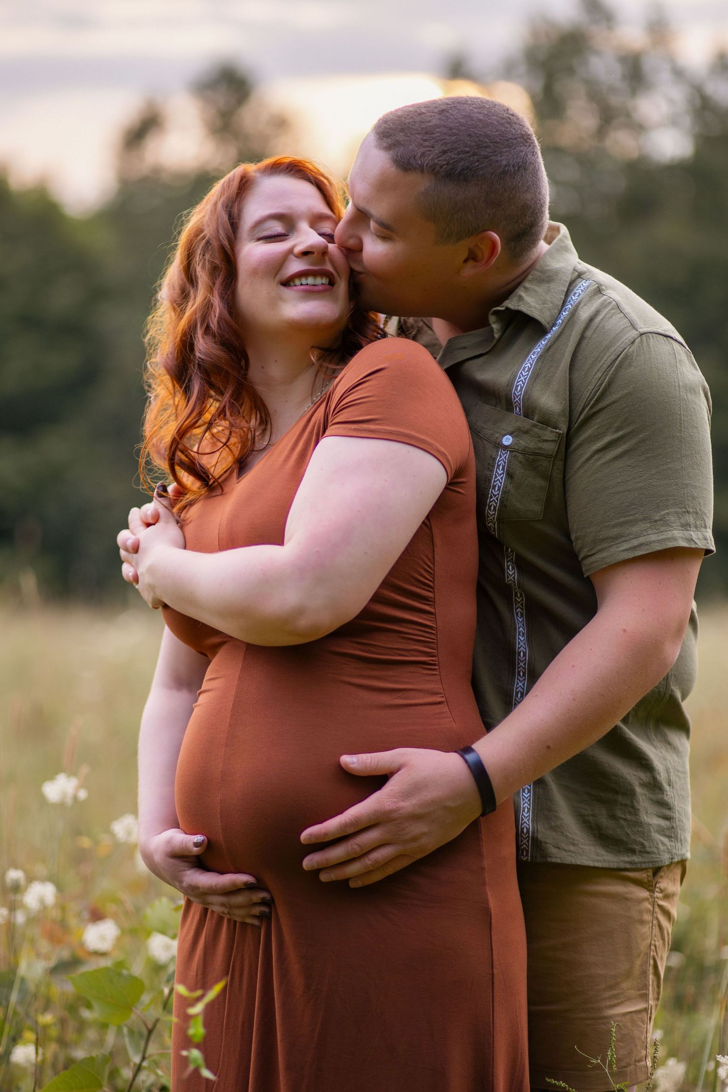 A romantic maternity photoshoot in a wildflower field at sunset shows a couple embracing tenderly.