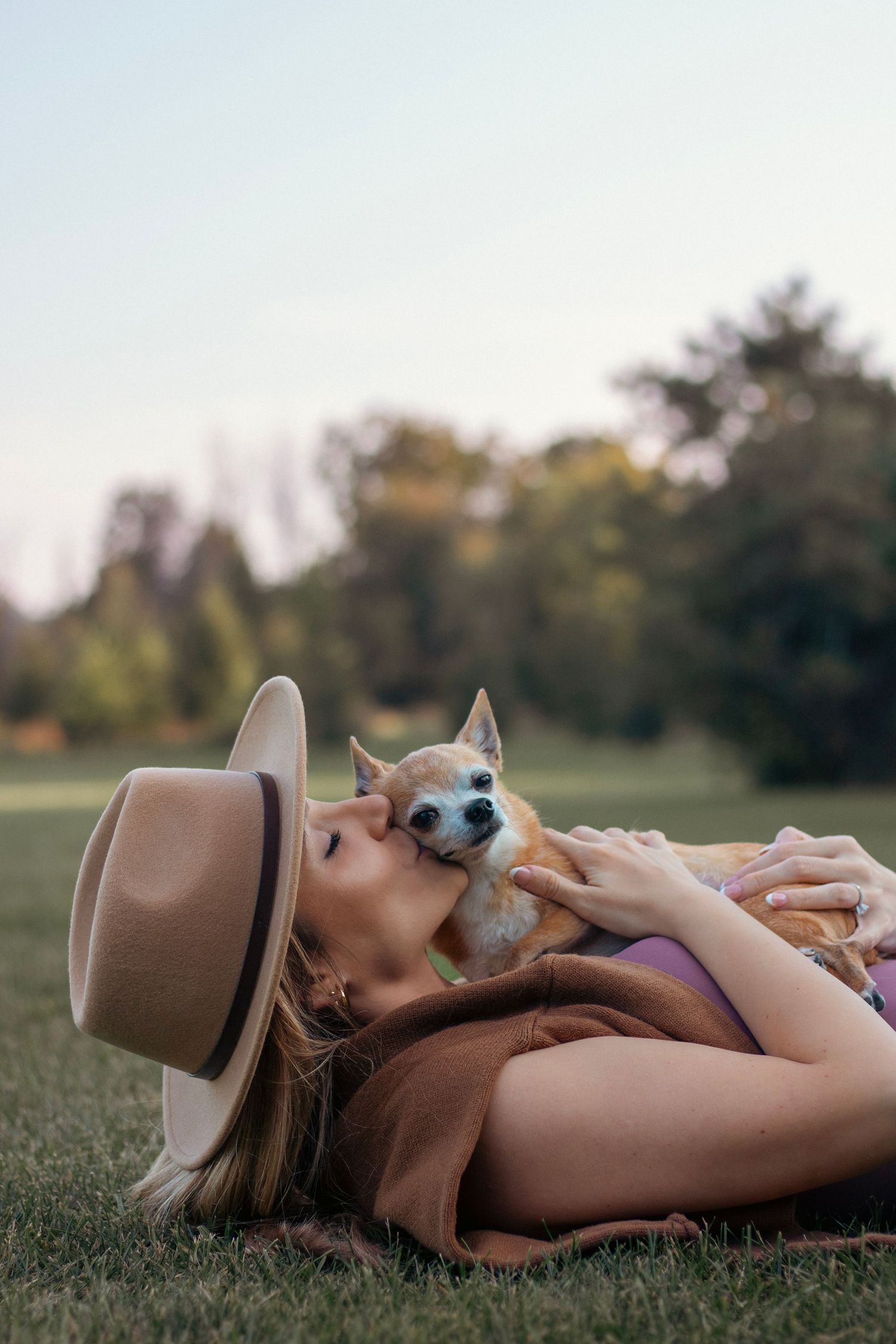 Person wearing a brown hat lies in grass with a small dog giving kisses.